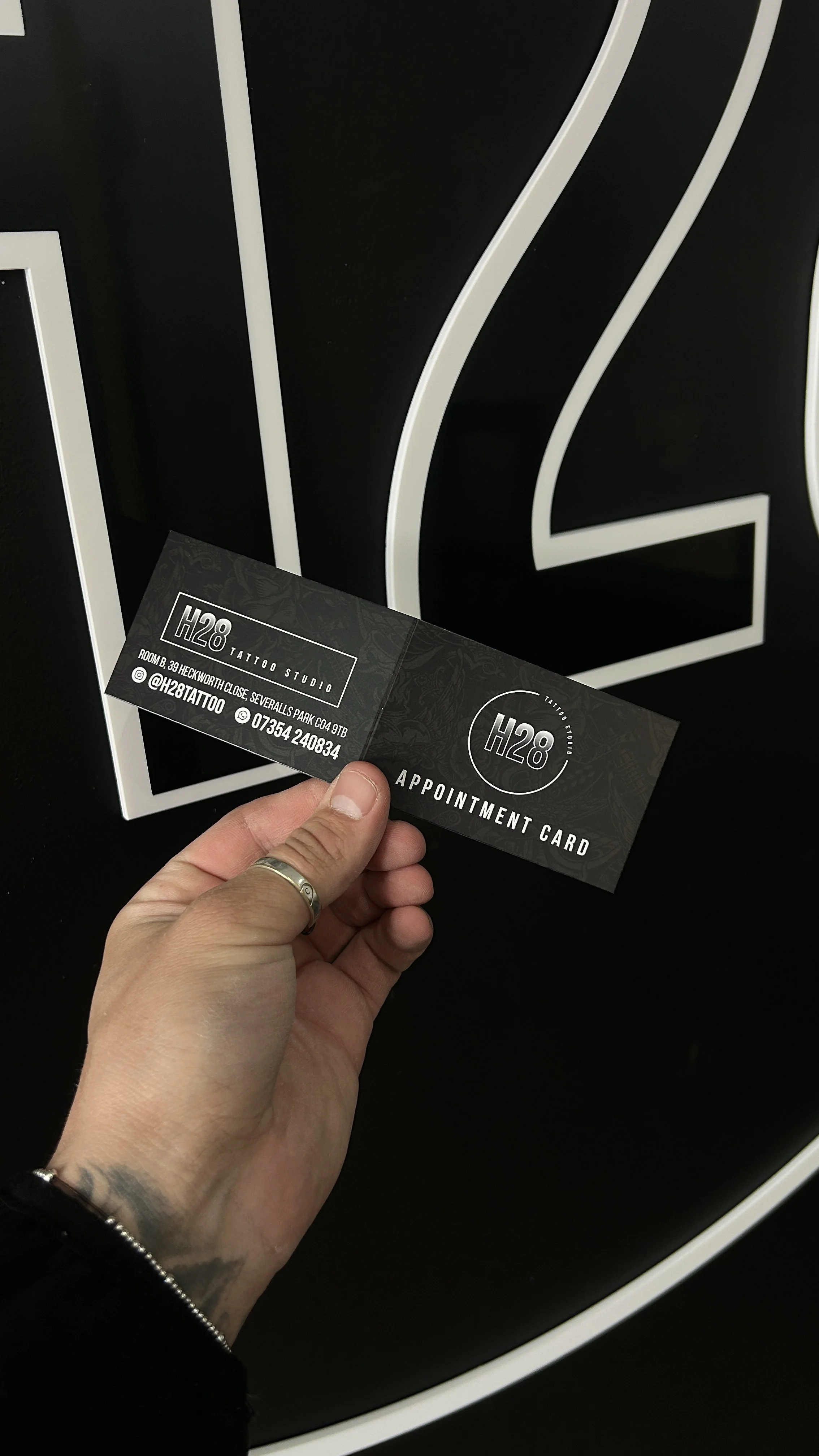 A person's hand holding a black appointment card for H28 Tattoo Studio in front of a large black and white geometric background.