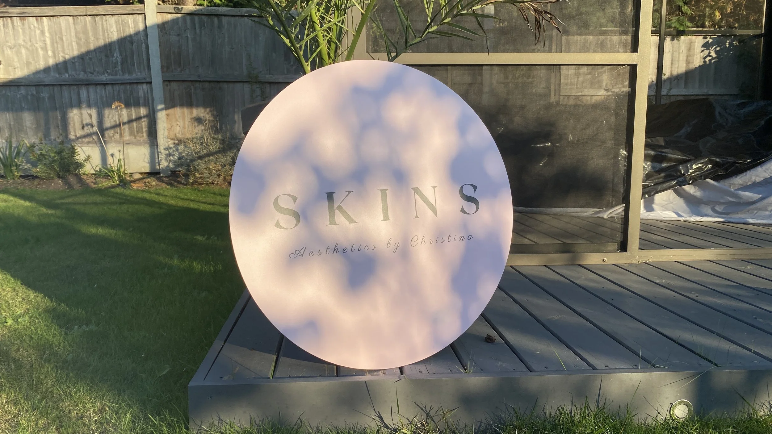 A circular sign with the word 'SKINS' and the phrase 'Aesthetics by Christina' in cursive, placed outdoors on a grassy lawn in front of a wooden fence and a greenhouse structure.