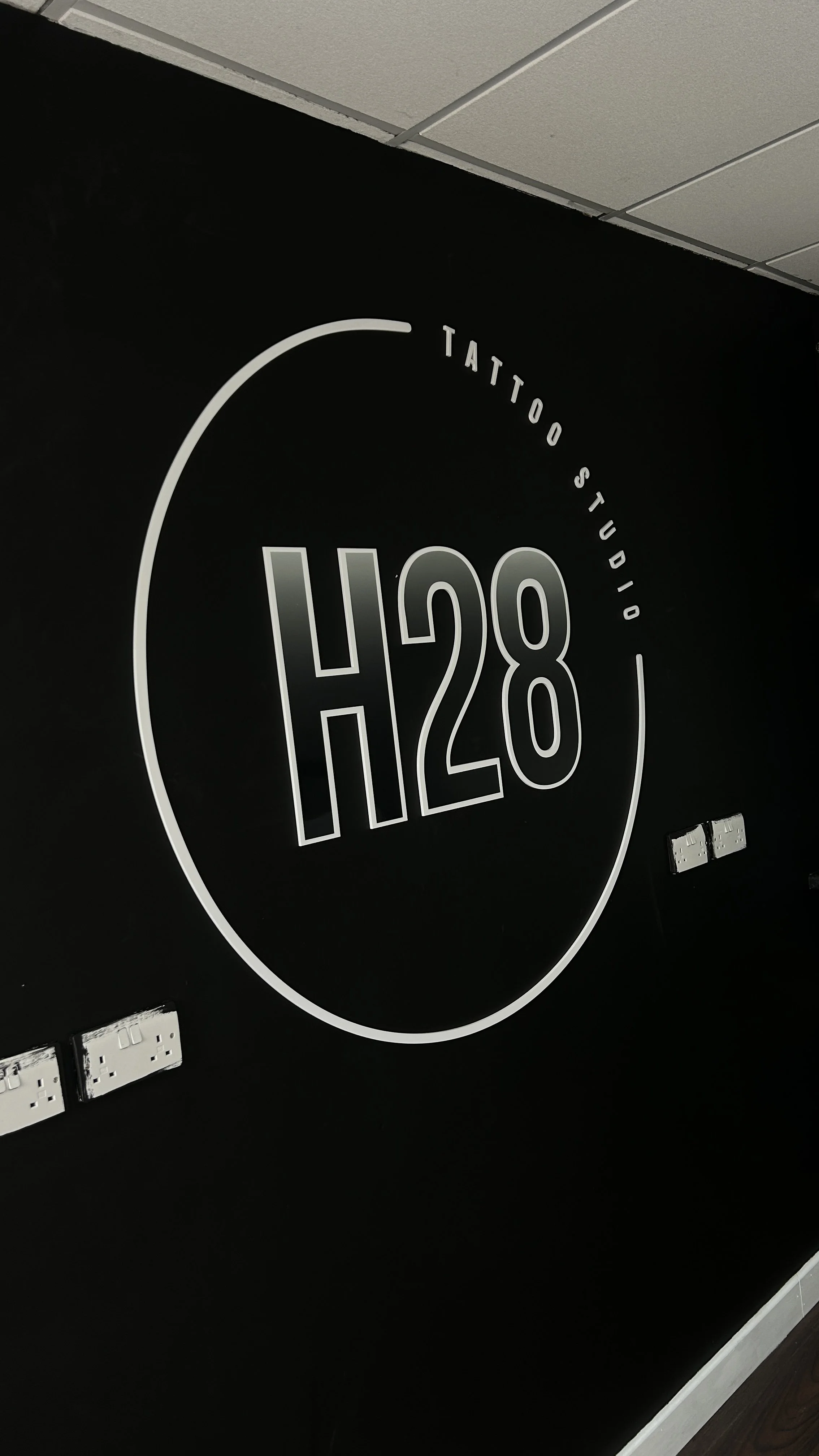 A black wall with a circular logo that reads 'H28' and 'Tattoo Studio' around it, with electrical outlets and cords visible.