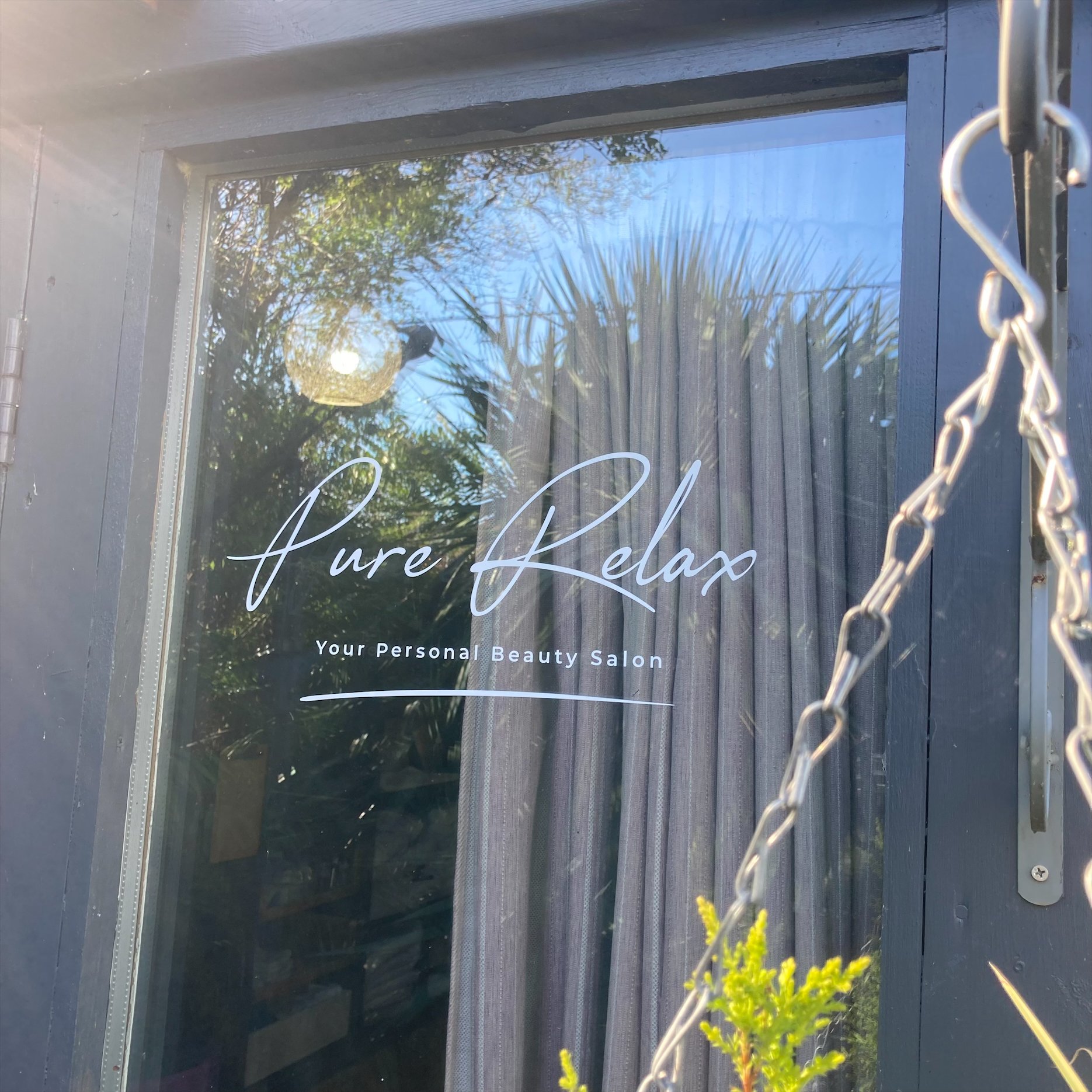 A window with a reflection of trees and sky. The window has a sign that reads 'Pure Relax.' There are some plants in the foreground and a chain hanging from the side of the window.