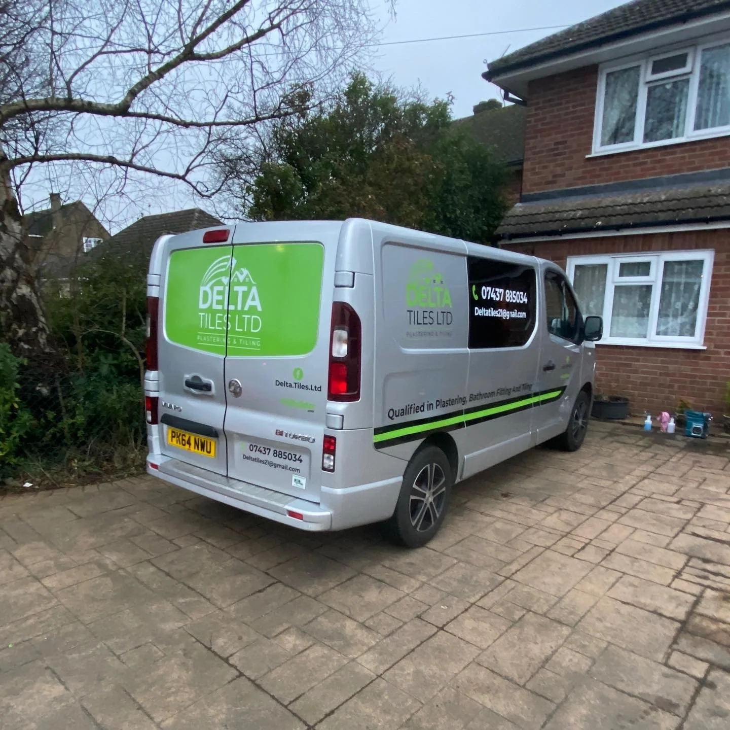A silver company van parked in front of a brick house with a window and some children's toys on the ground. The van has green and black branding for Delta Tiles Ltd, a phone number, email, and services listed, with a license plate number of PK64 NWU.