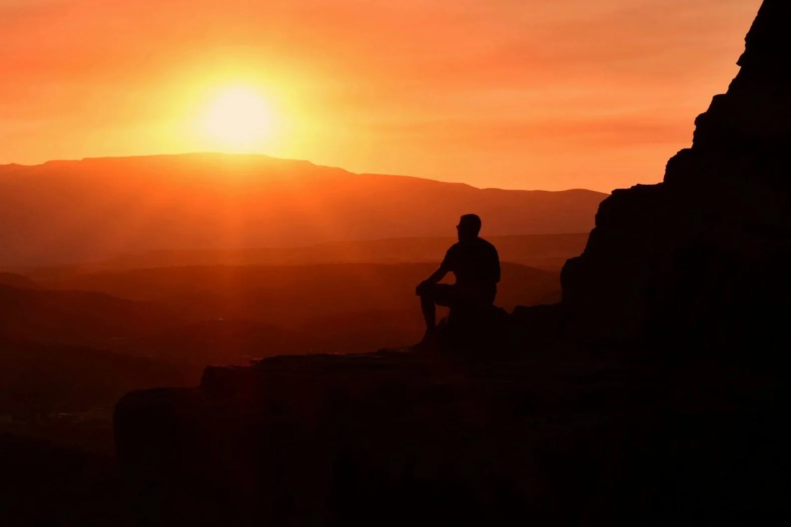 Silhouette of a person sitting on a rock at sunset, with a view of mountains in the background and a glowing sun in the sky.