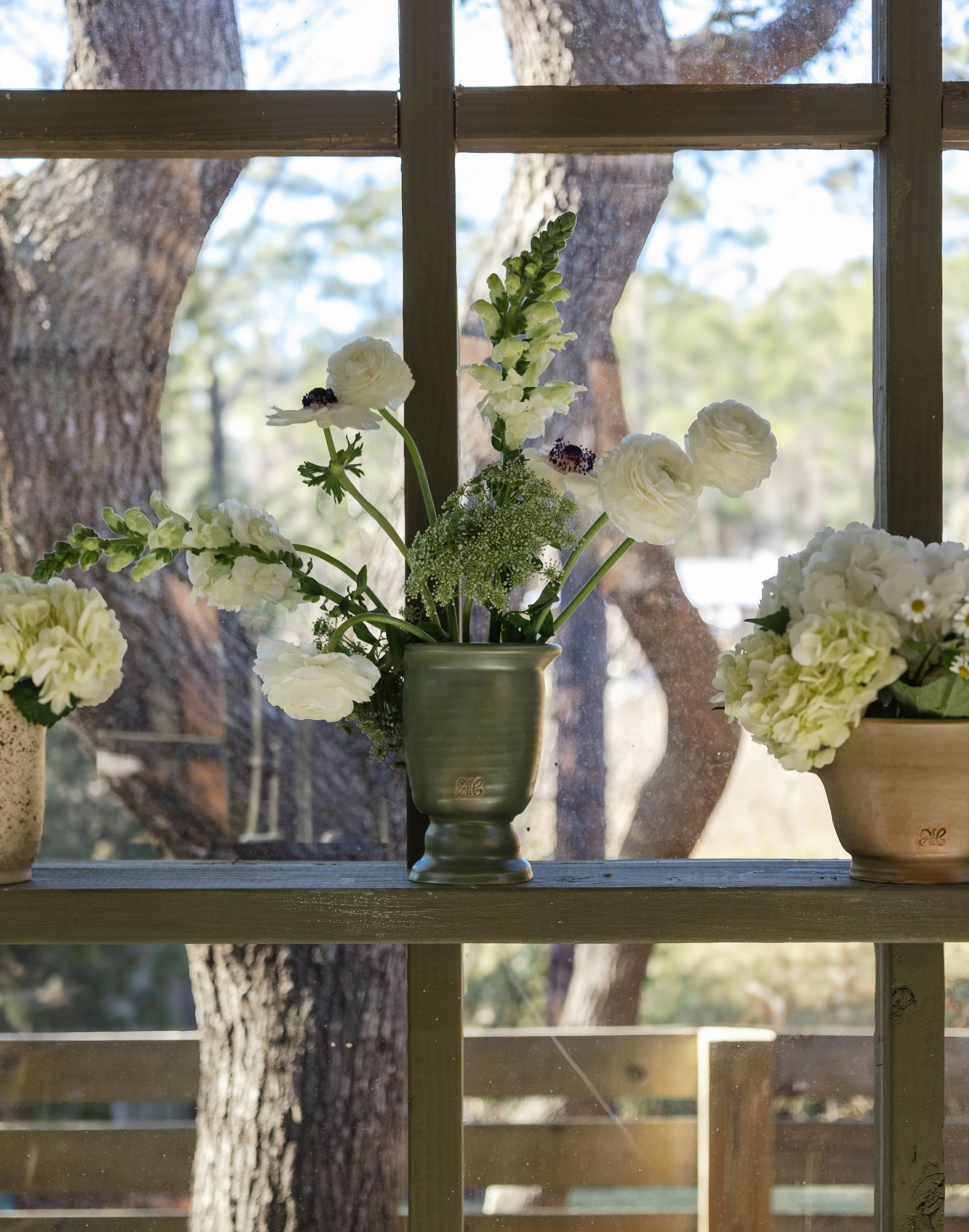 Vase with white flowers on a wooden shelf in front of a window with trees outside.