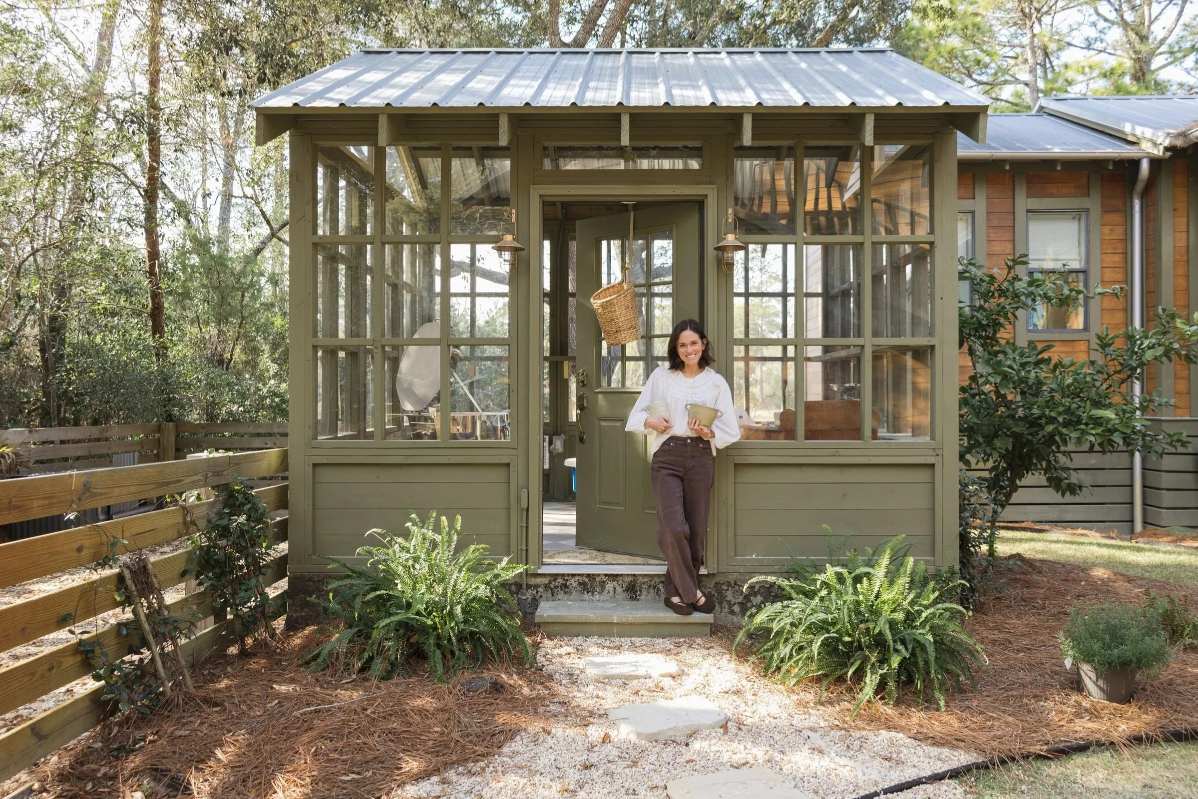 Woman standing in front of a small green porch with glass walls, holding a coffee mug and a bowl, smiling, in a backyard with plants and trees.