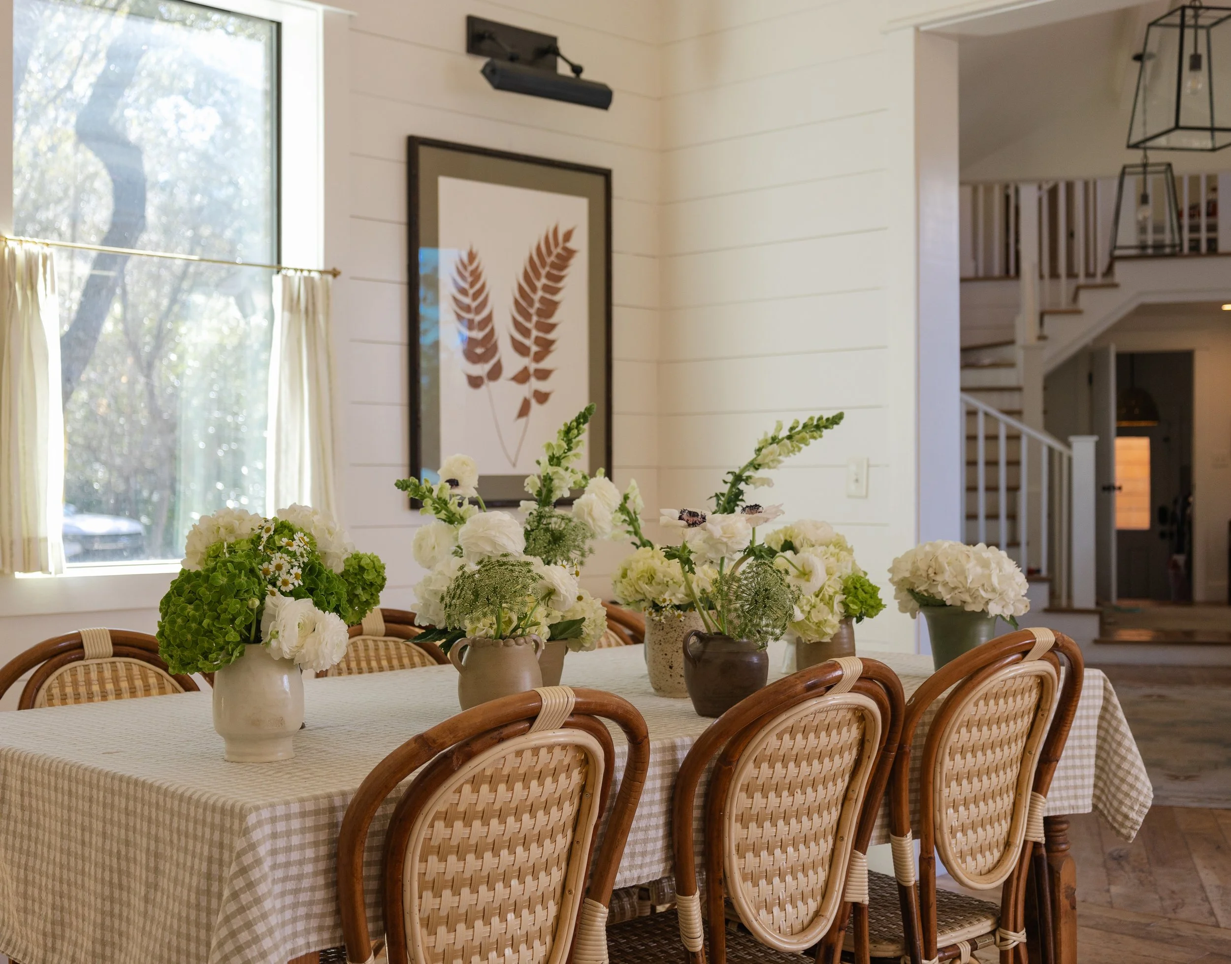 A dining table with a checkered tablecloth and six wooden chairs with cane backs. The table is decorated with five vases filled with white and green flowers. A window with cream curtains is in the background, along with framed artwork of two brown le