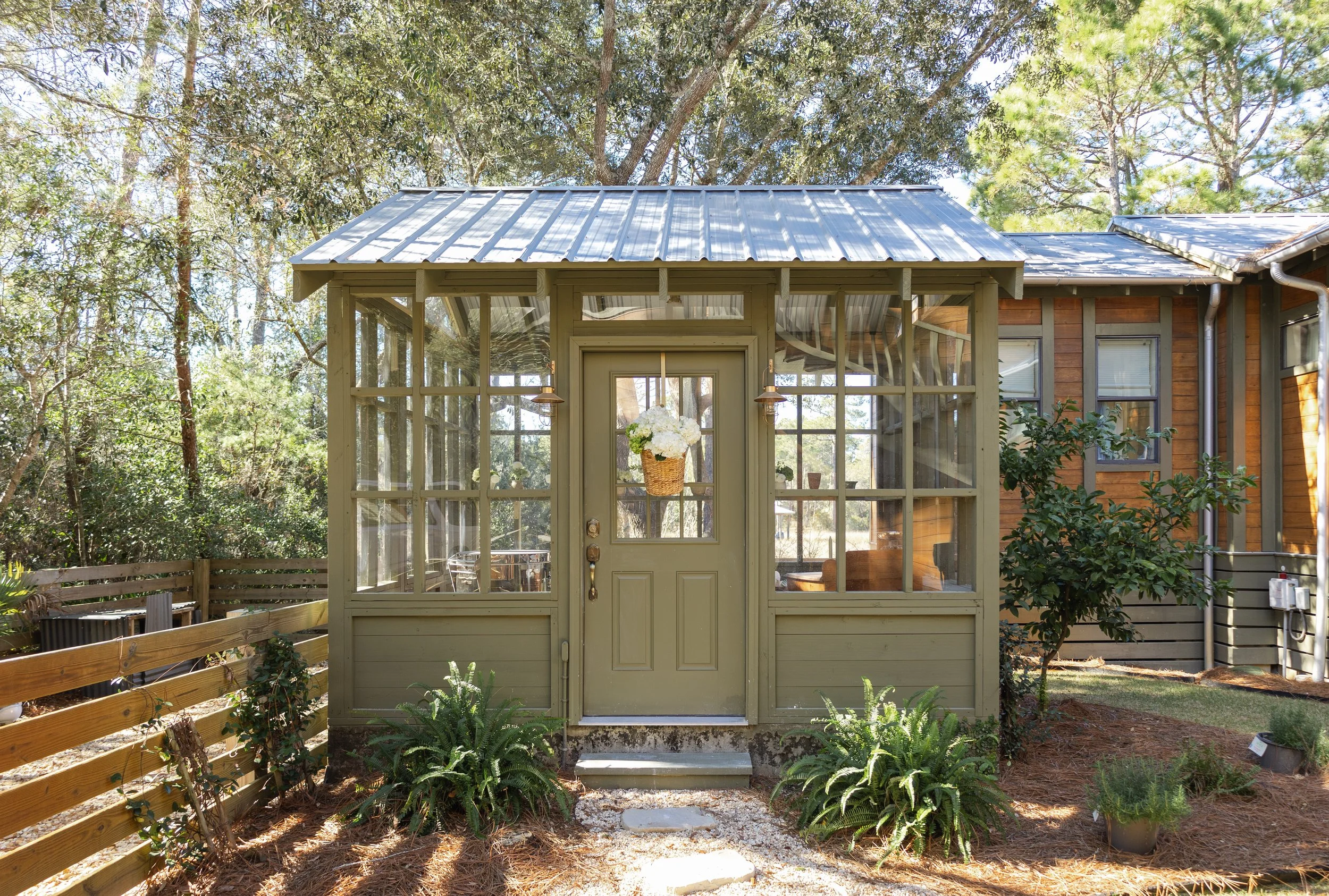 Green backyard sunroom with glass windows, vaulted metal roof, front door with hanging flower basket, and surrounding plants and trees.