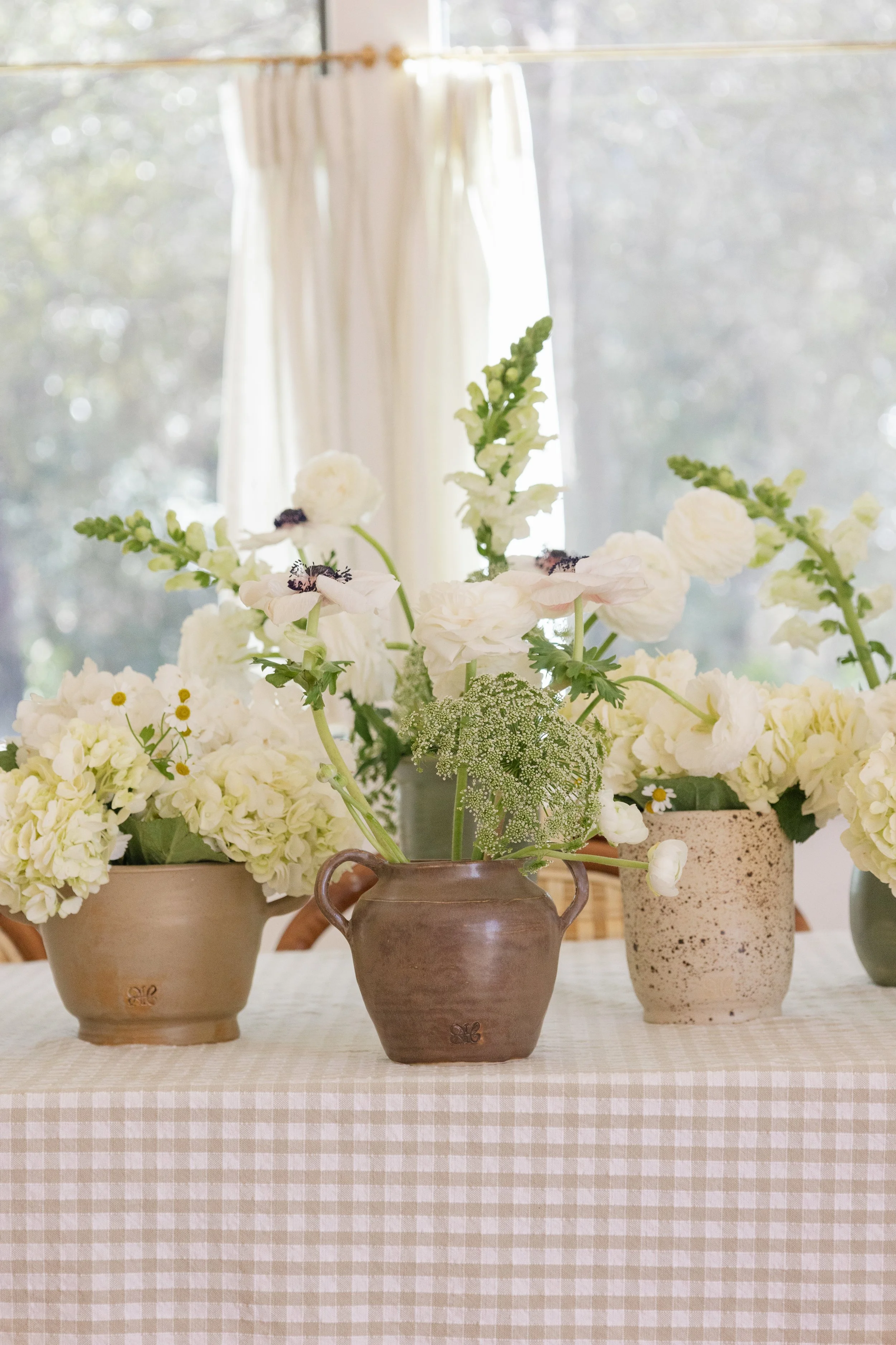 Vases with white flowers on a checkered tablecloth in front of a window with cream curtains.