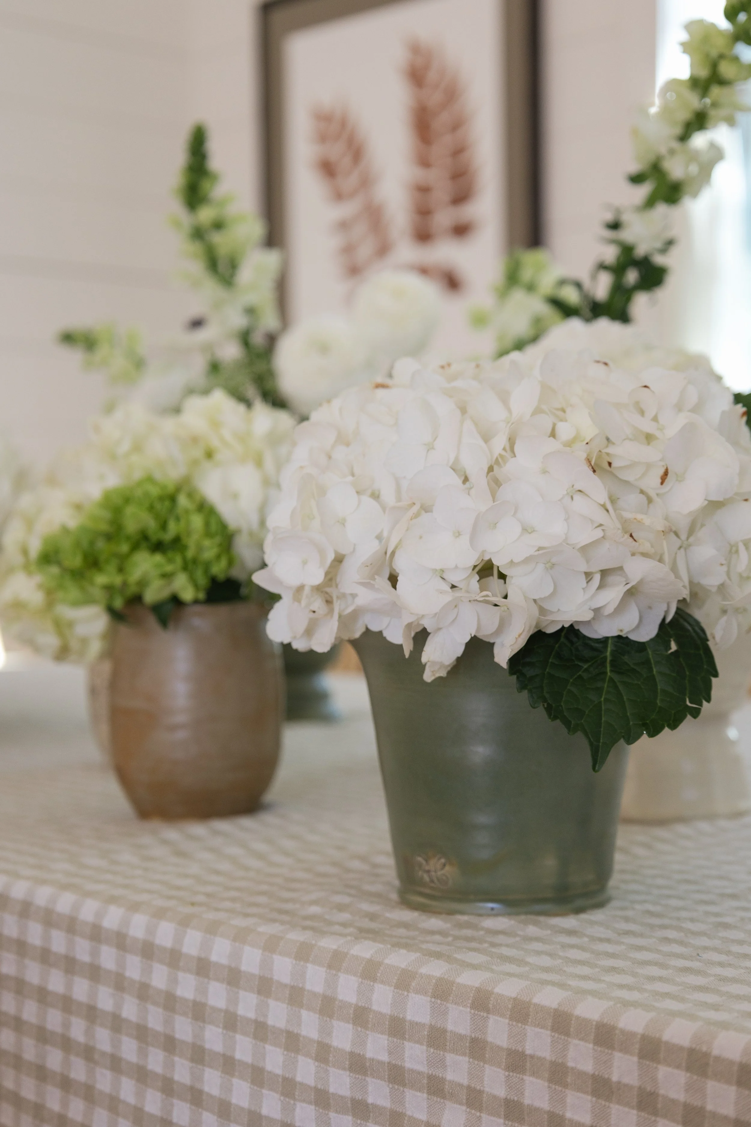Close-up of a white hydrangea flower arrangement in a ceramic green pot on a checkered tablecloth, with other similar flower arrangements and a framed botanical print in the background.