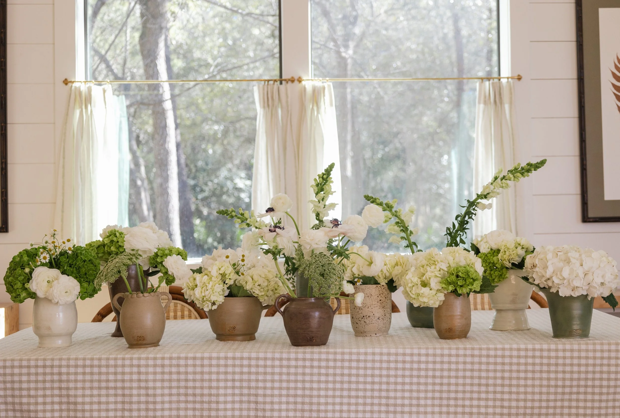 A table with various white flower arrangements in different vases, set in front of a window with light-colored curtains and a wooded view outside.
