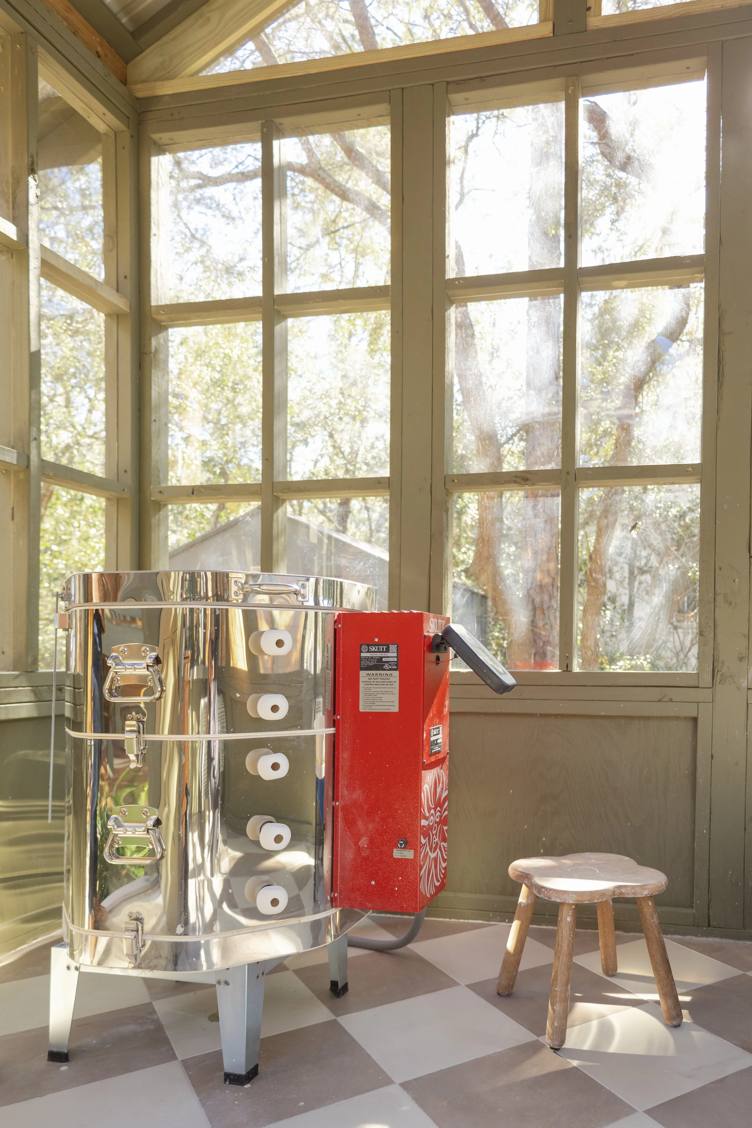 A shiny silver cylindrical pizza oven with a red control box attached, standing on four legs, in a sunlit room with large windows. There is a small, rustic wooden stool nearby and a checkered floor.