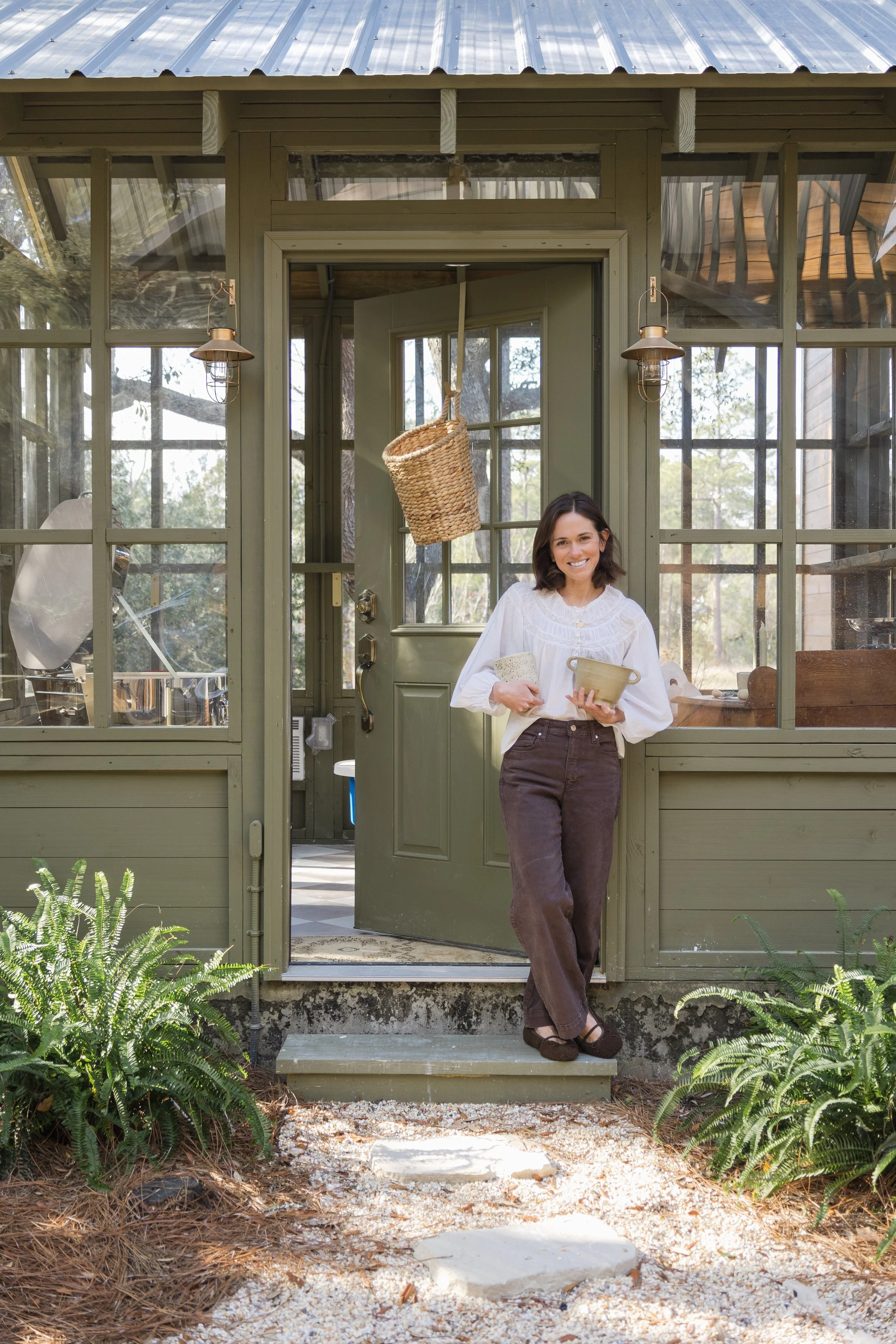 A woman standing at the entrance of a green greenhouse, holding a collection of gardening tools, smiling, with plants and gardening supplies visible inside.