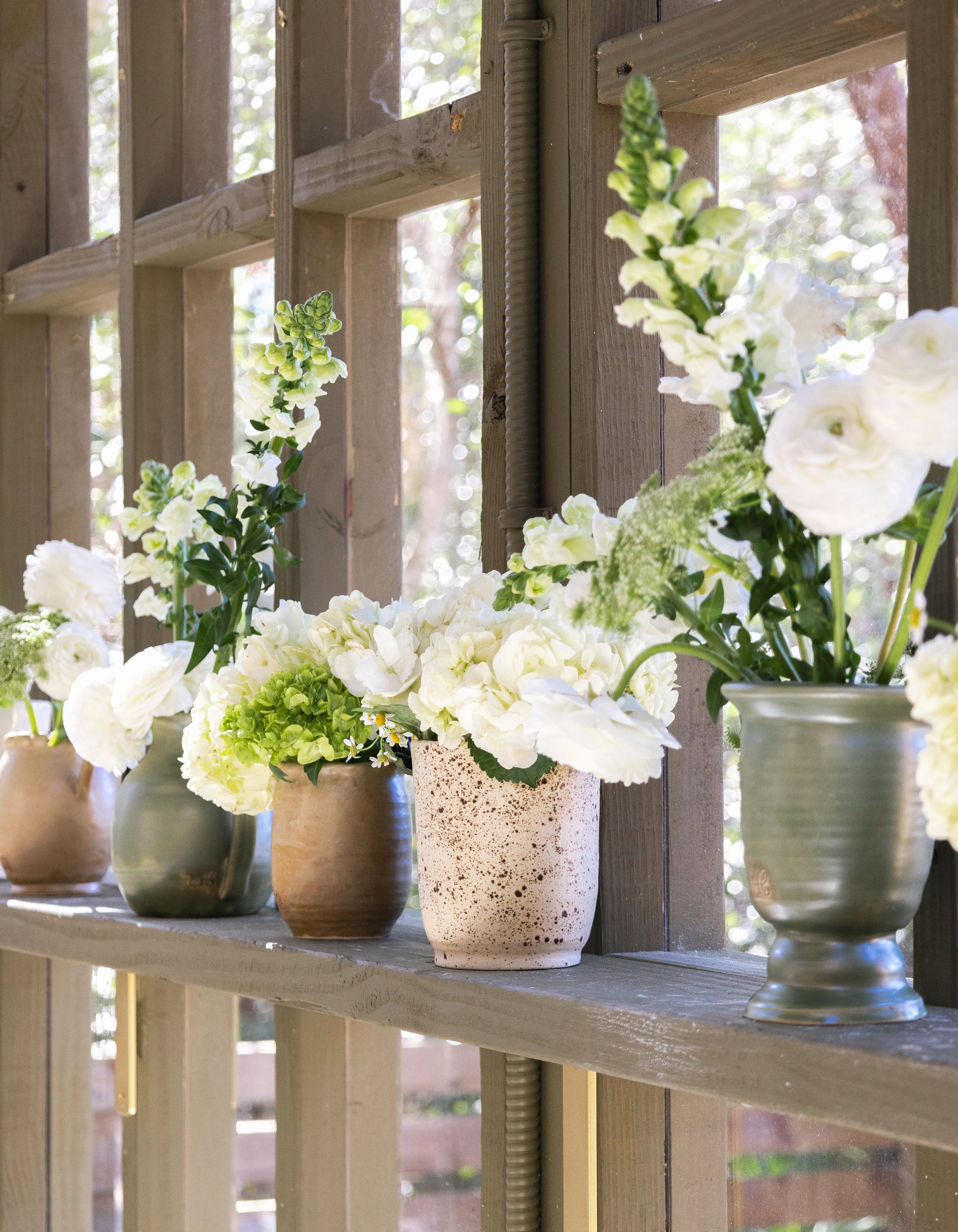 A row of vases filled with white flowers on a wooden shelf, with a window and outdoor trees in the background.