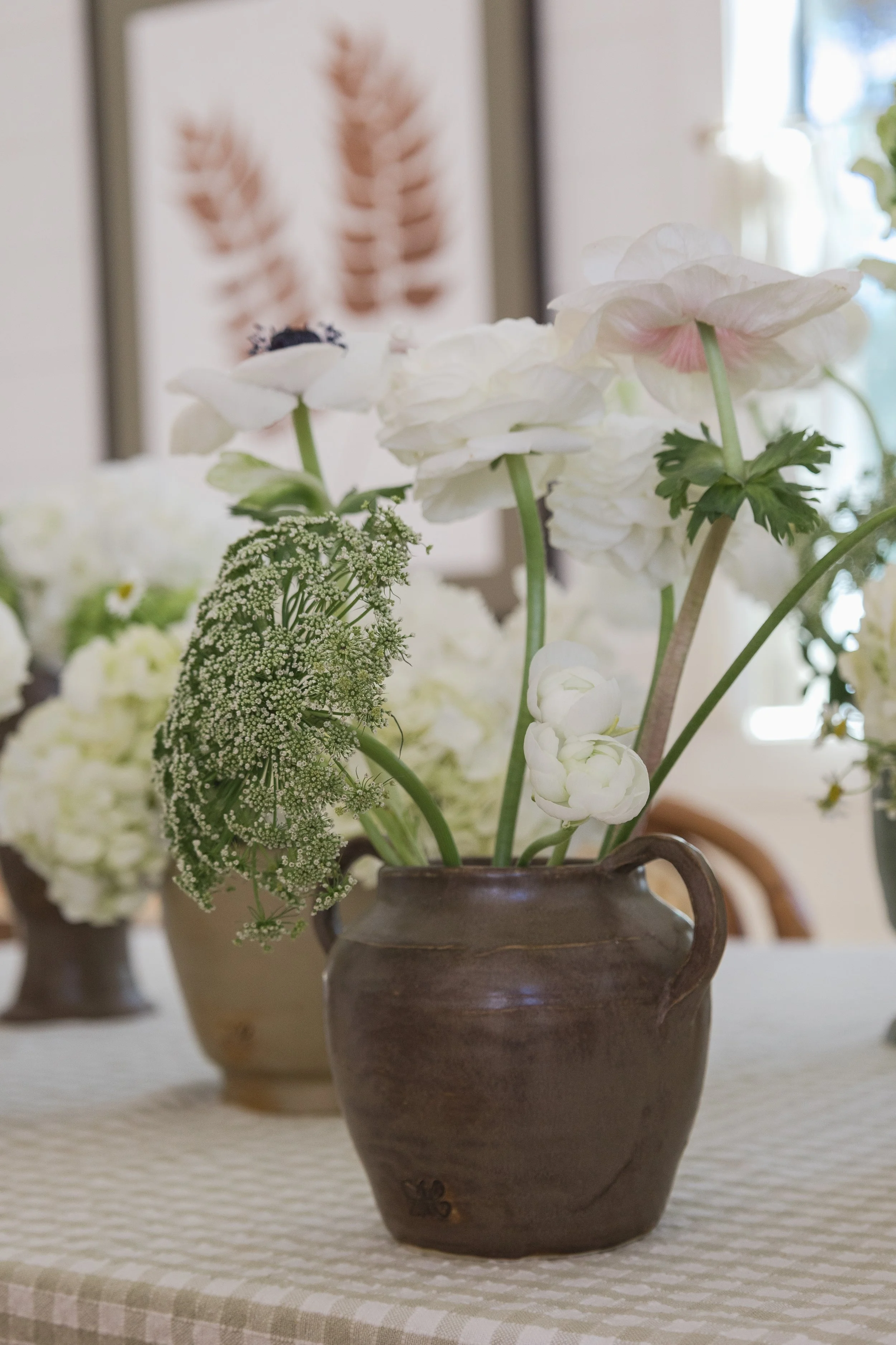 A brown ceramic vase filled with white garden flowers, including ranunculus and Queen Anne's lace, on a table with a checkered tablecloth, with framed botanical art in the background.