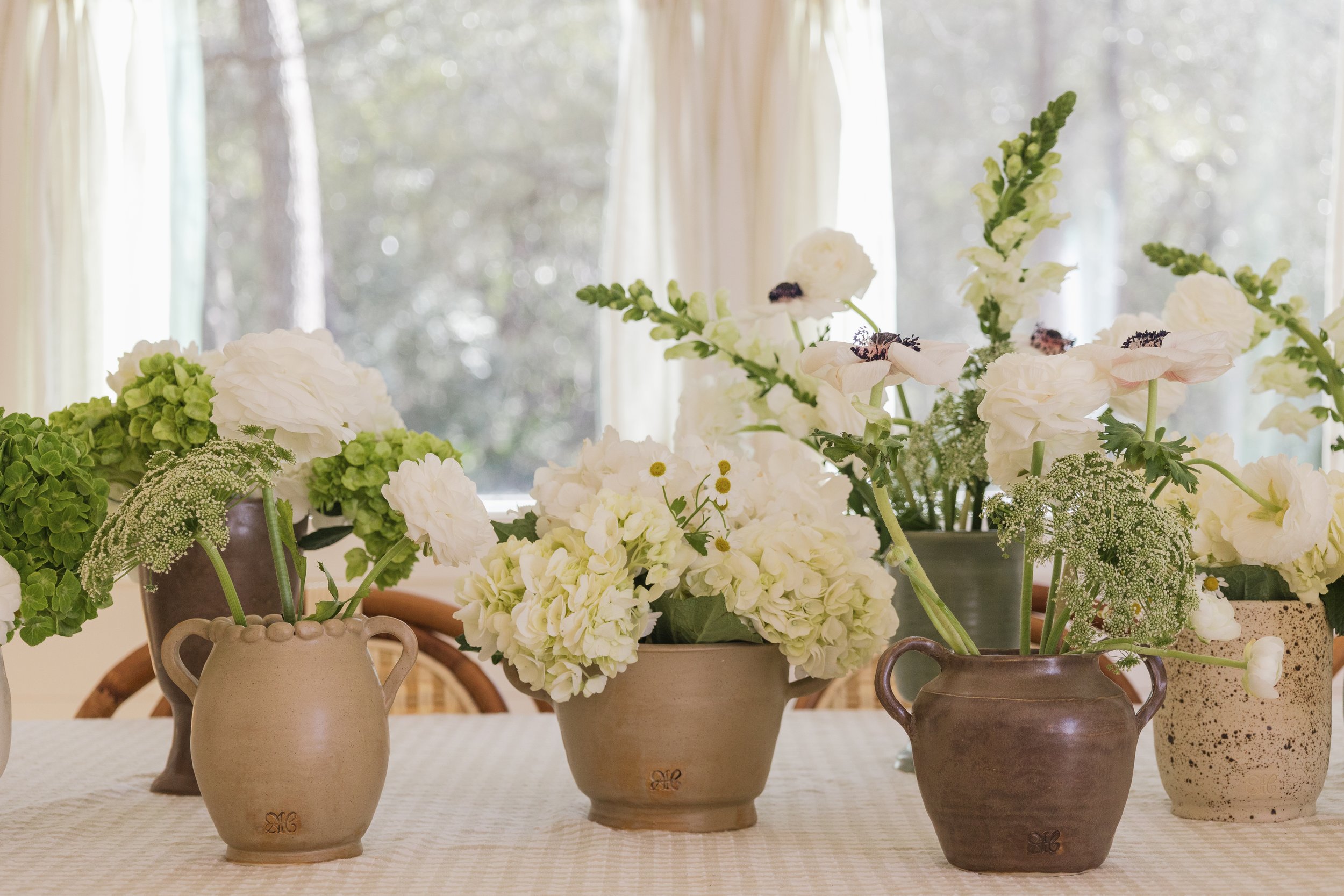 Arrangement of white and green flowers in various vases on a table with a blurred window in the background.