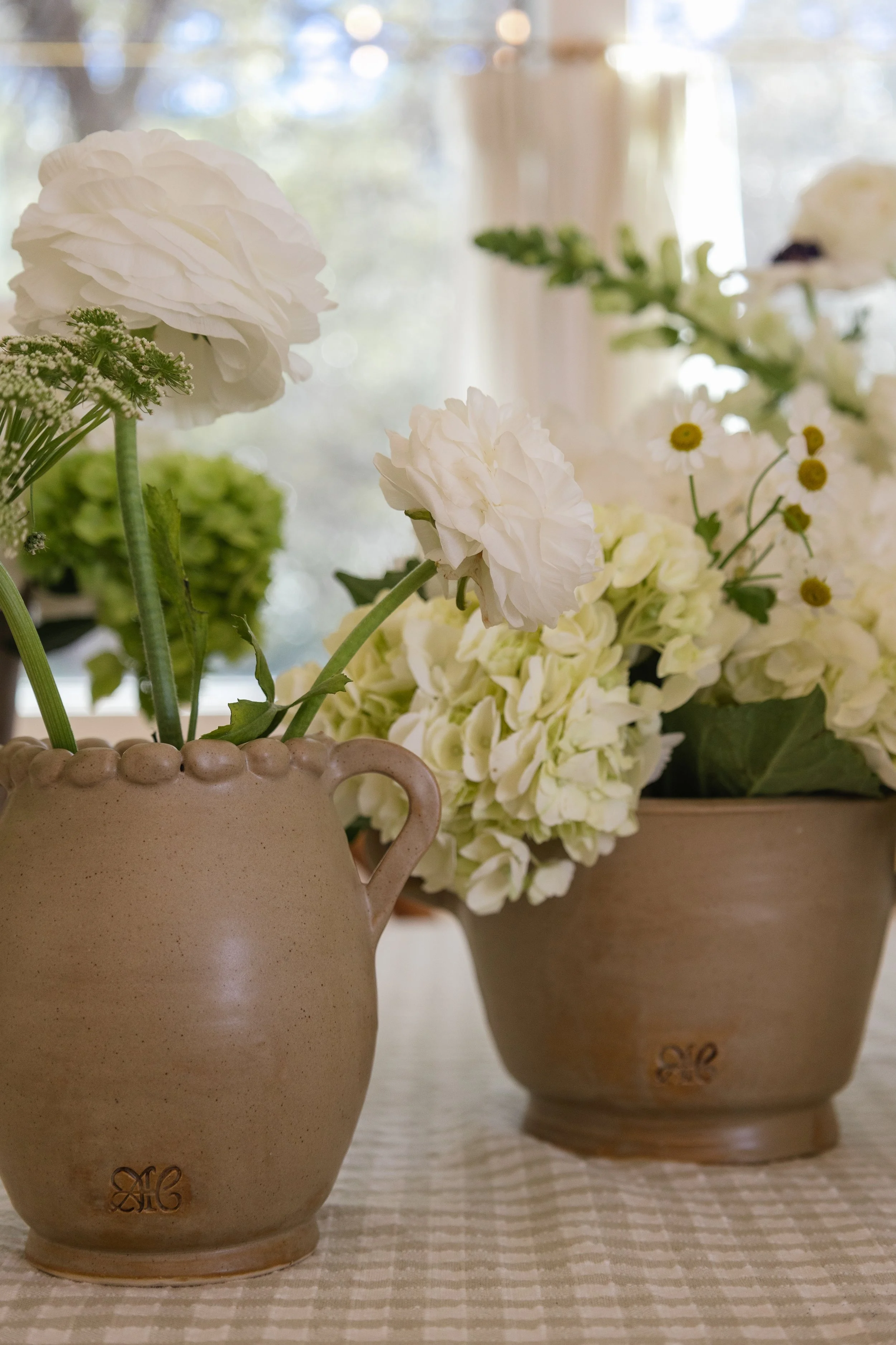 Two beige vases with white flowers and greenery on a checkered tablecloth, with a blurred window background.