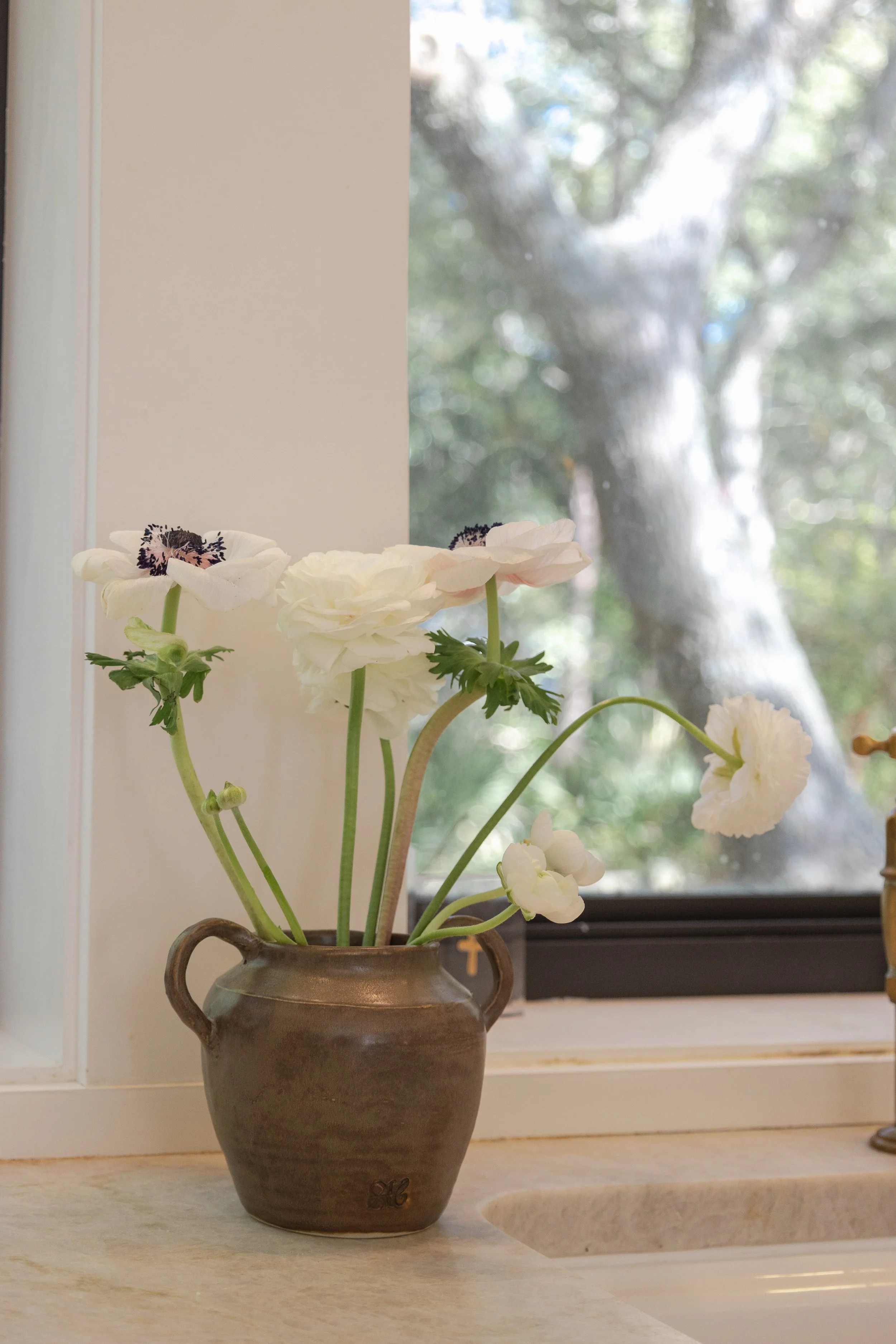 A ceramic vase with white flowers on a kitchen countertop near a window with a view of trees outside.