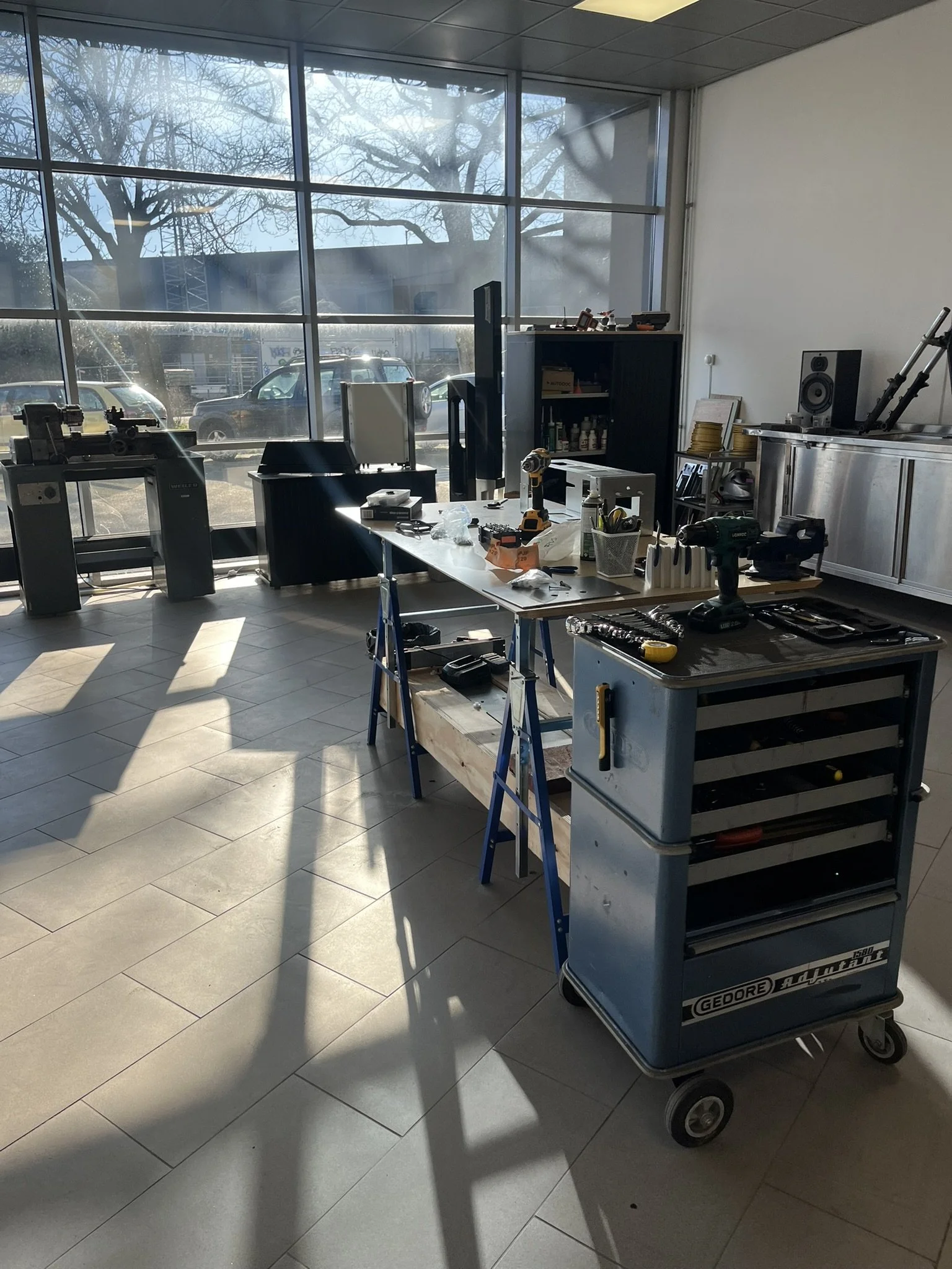 An interior office or workshop space with large front windows showing parked cars and bare trees outside. The room contains work tables with tools, supplies, and equipment, including a work cart, tools, and a cabinet.