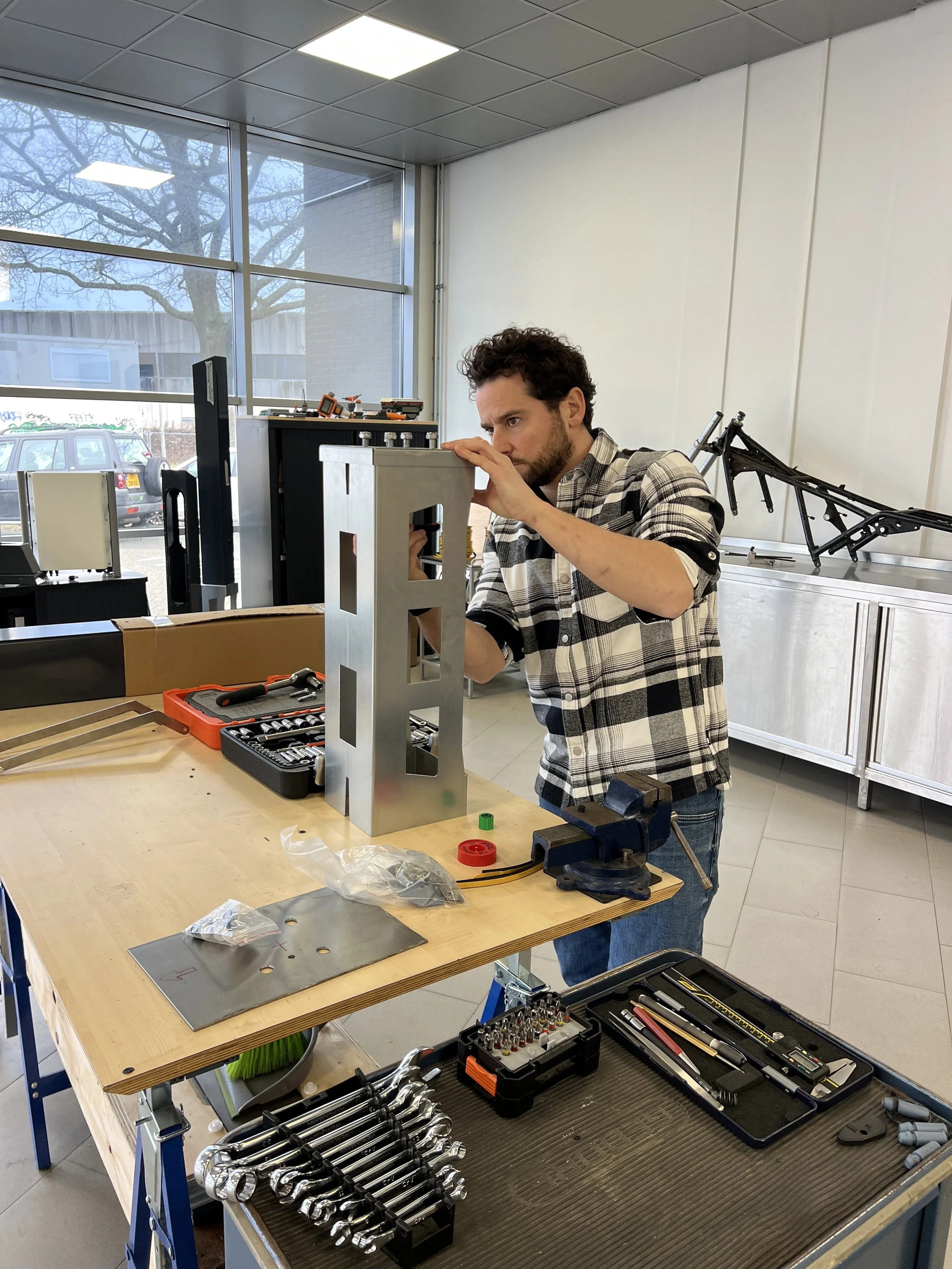 Man working on mechanical assembly at a workbench inside a workshop with tools and parts, some bicycle frames are visible in the background.