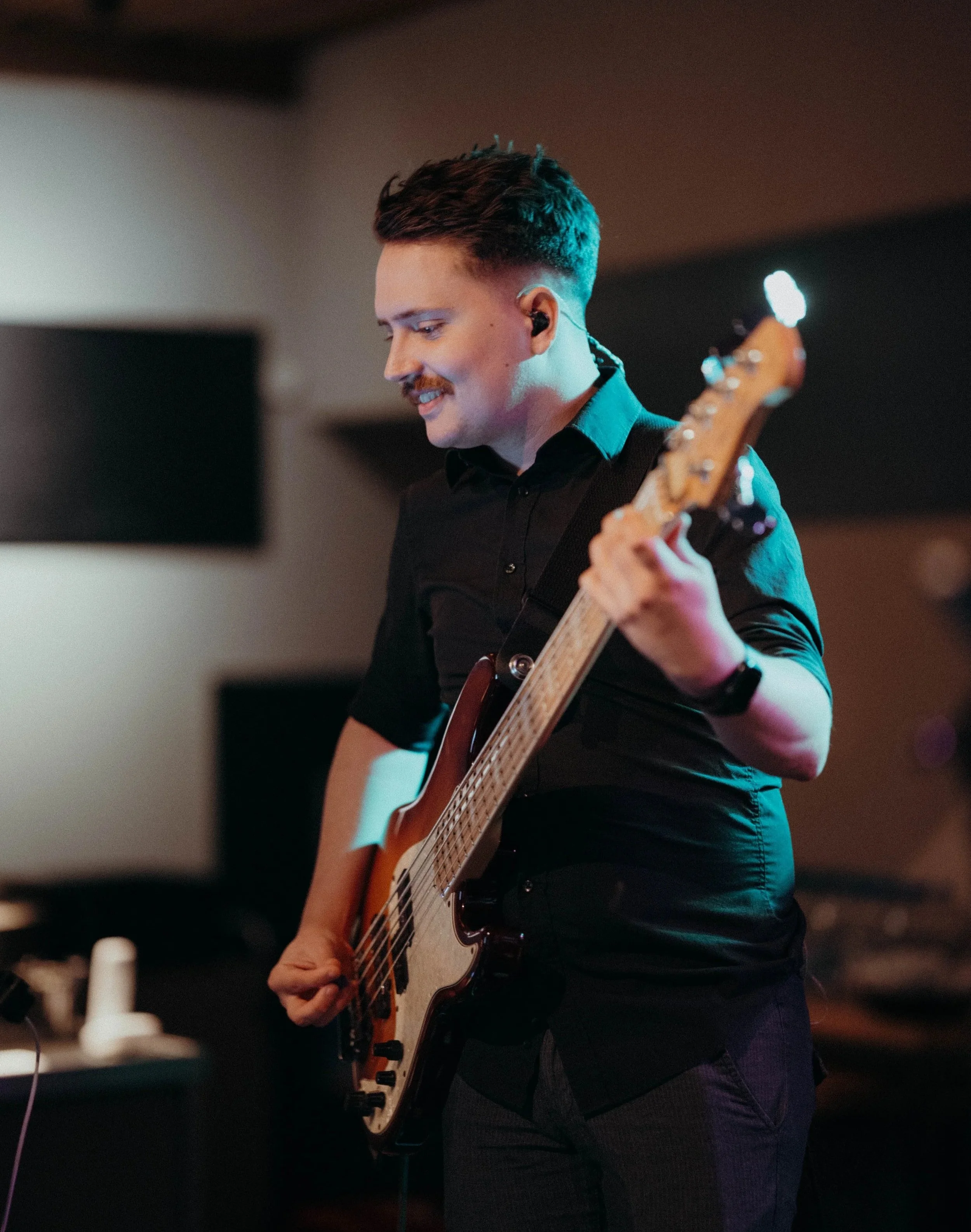 Ein junger Mann spielt E-Bass auf einer Bühne, trägt eine schwarze Hemd und hat einen Seitenscheitel. Er lächelt und sieht konzentriert aus, während er spielt.