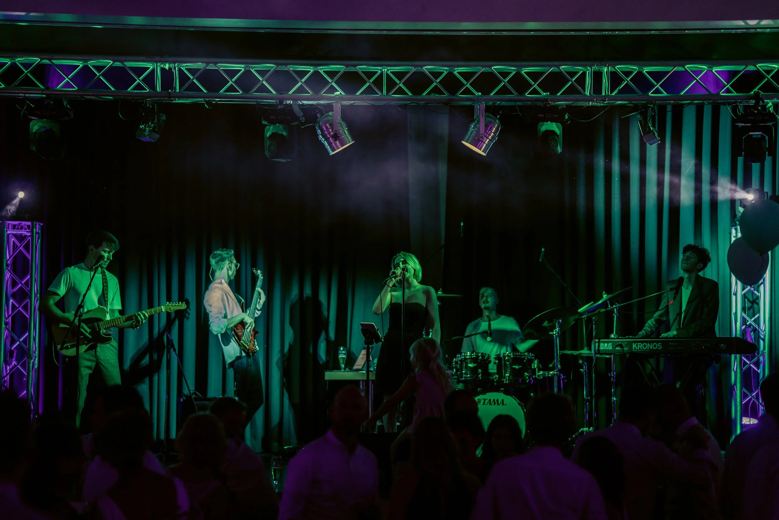 Eine Band tritt bei einem Konzert auf einer Bühne mit dunklen Vorhängen auf, beleuchtet von bunten Scheinwerfern, während das Publikum im Vordergrund feiert und tanzt.