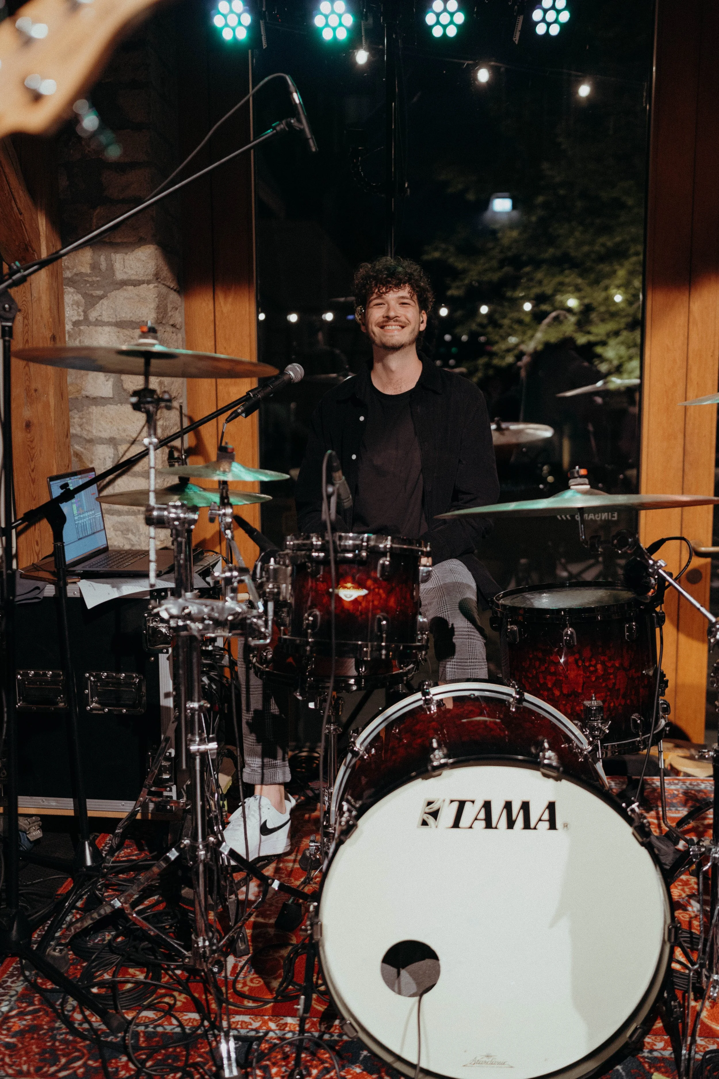 Ein lachender junger Mann sitzt hinter einer roten TAMA Schlagzeug. Er trägt eine schwarze Jacke und schwarze T-Shirt. Es ist auf der Bühne mit der Band casual chic mit Holzrahmen und großen Fenstern im Hintergrund.