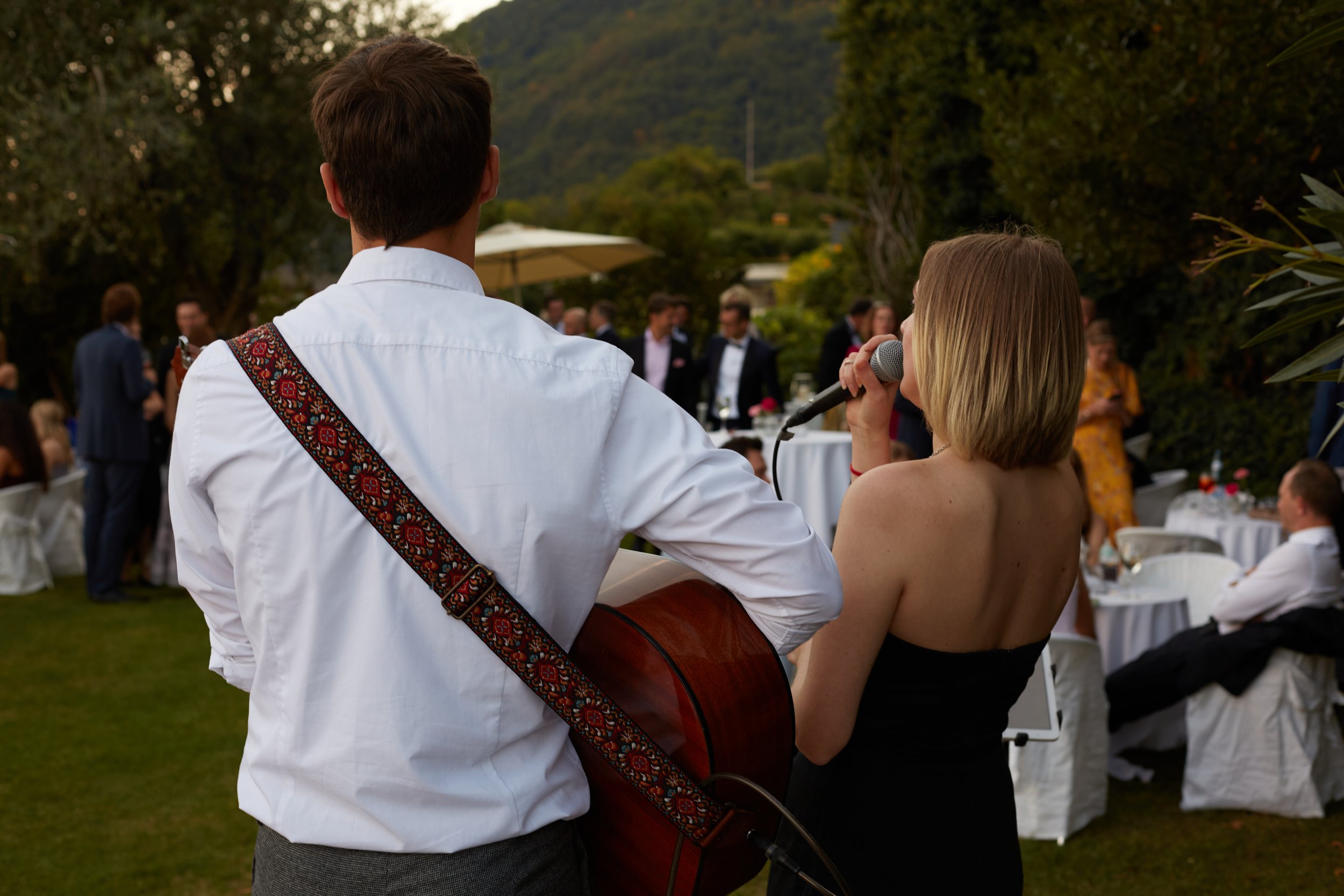Bei einer weißen Hochzeit in Italien spielt ein Mann mit Akustikgitarre und toller Hochzeitssängerin Livemusik für die Gäste im Garten.