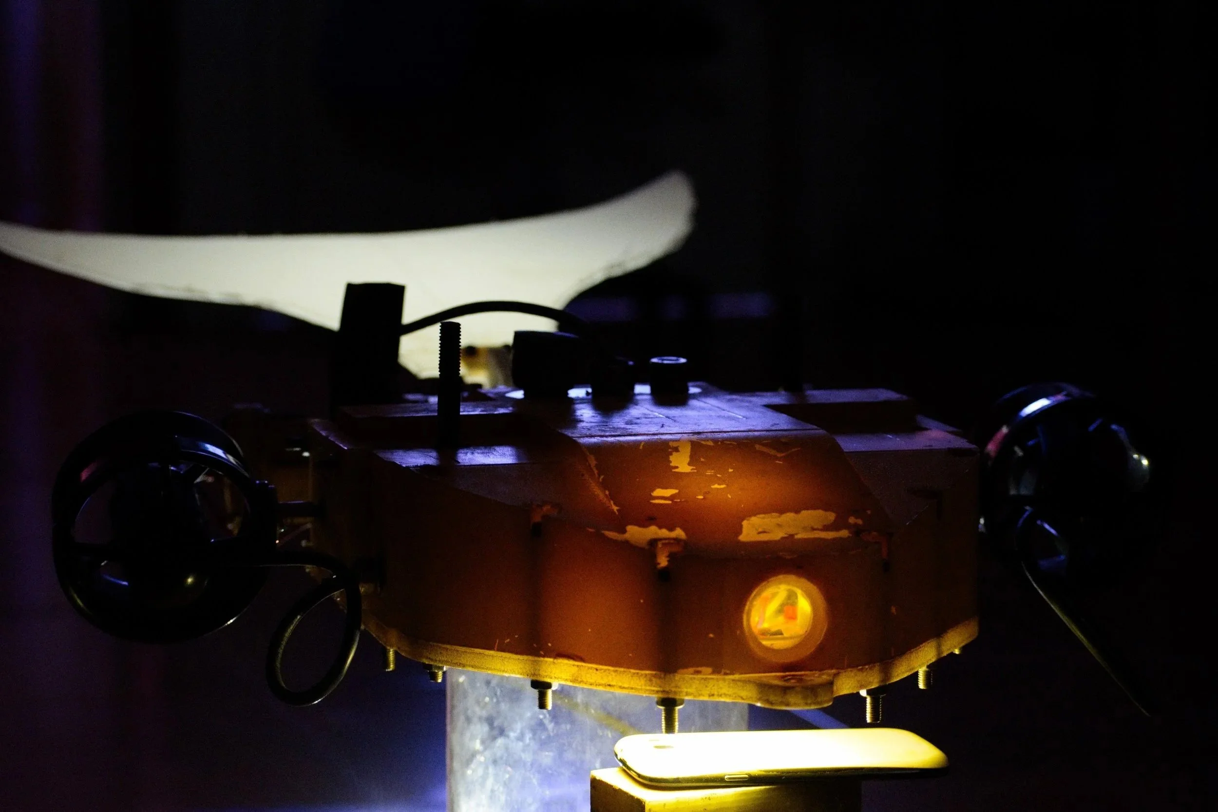 Close-up of a small drone or robotic device with two propellers, illuminated by a yellow light, in a dark environment with a large, white, crescent shape in the background.