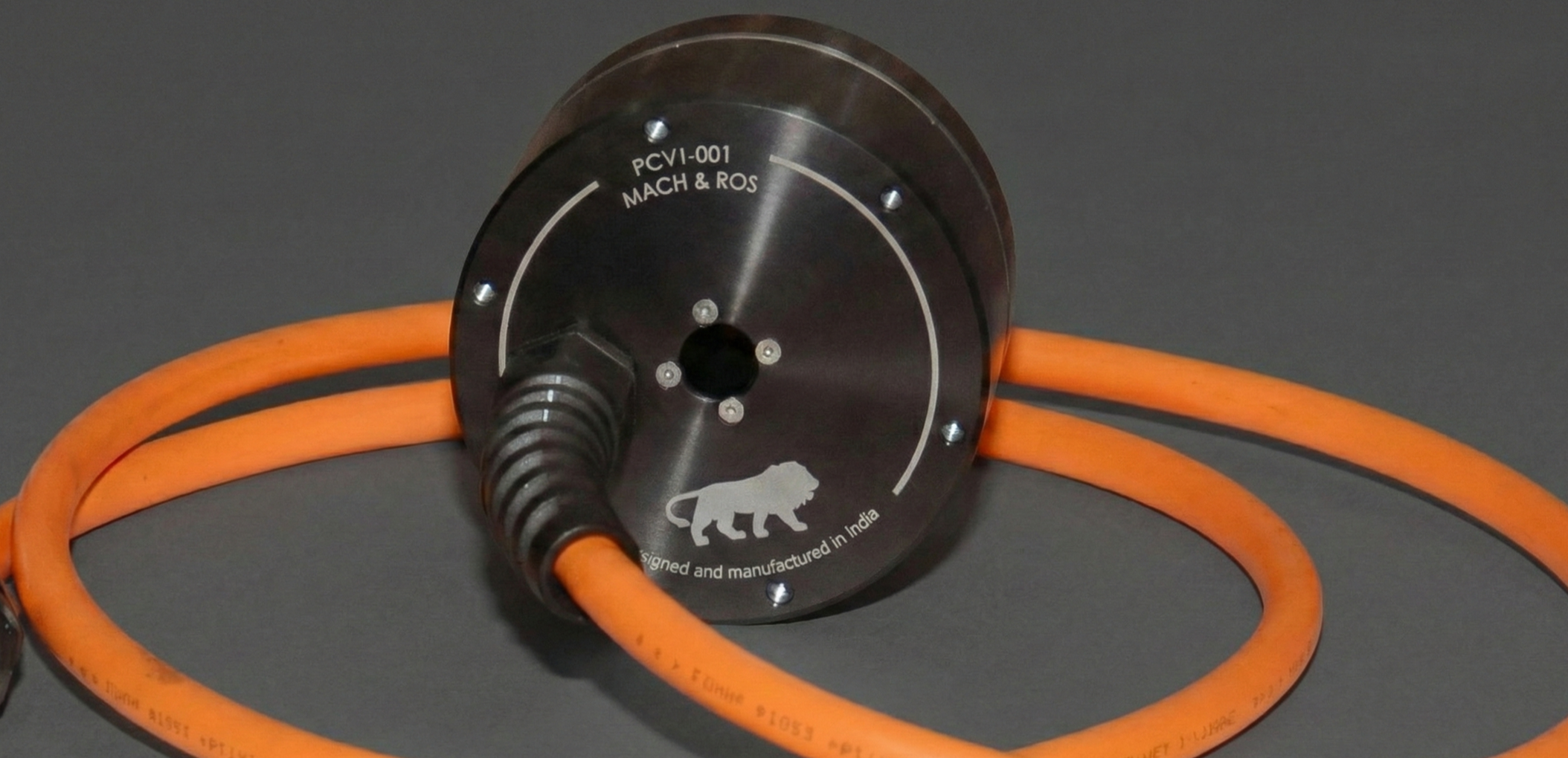 Close-up of a cylindrical metal device with orange cables attached, featuring a lion logo and text indicating it was designed and manufactured in India; the device has labels including PCVI-001 and Mach & ROS.