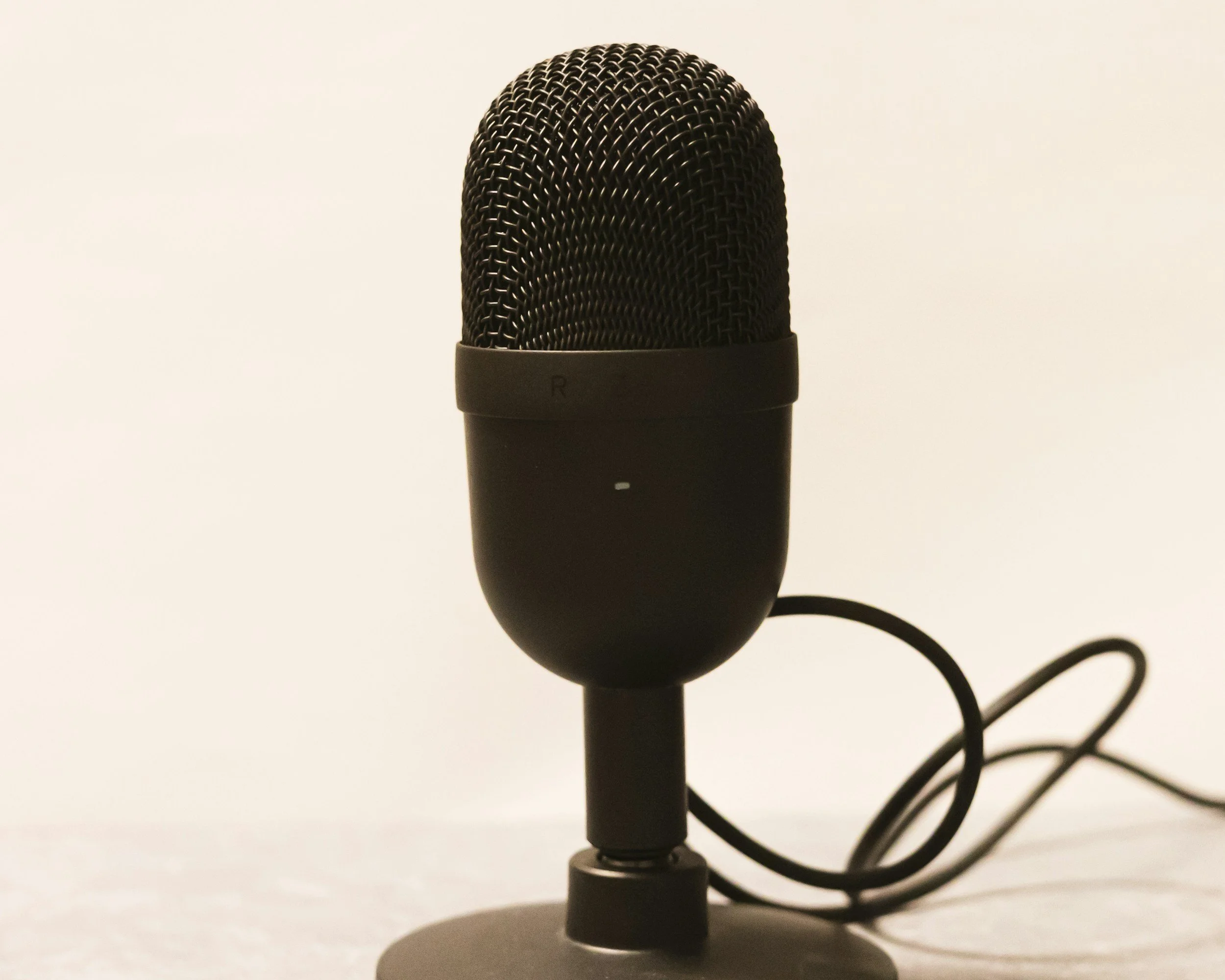 A black studio microphone with a mesh grille on a stand, connected by a cable.