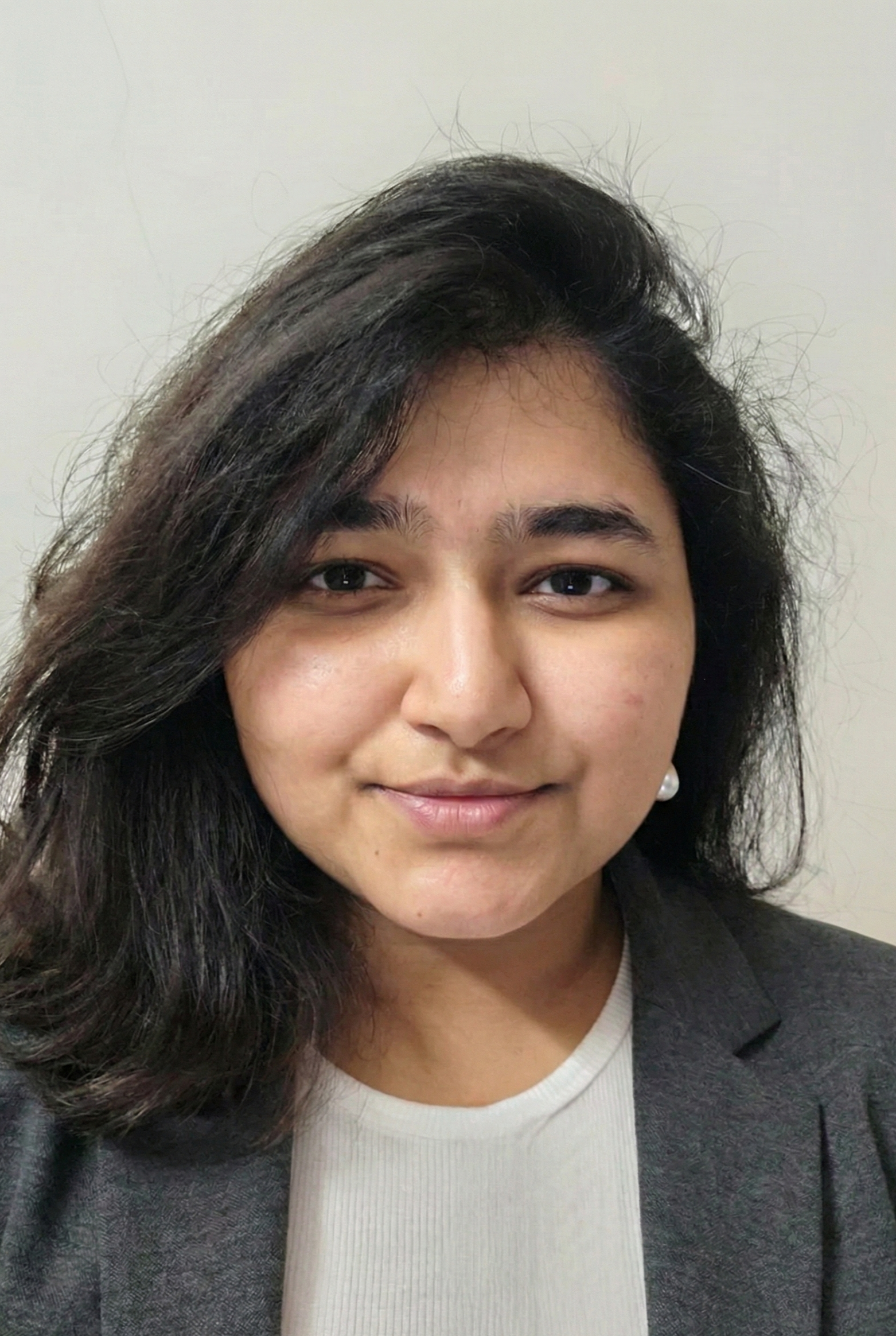 A young woman with dark, shoulder-length hair, wearing a gray blazer and a white top, smiling mildly against a plain light-colored background.