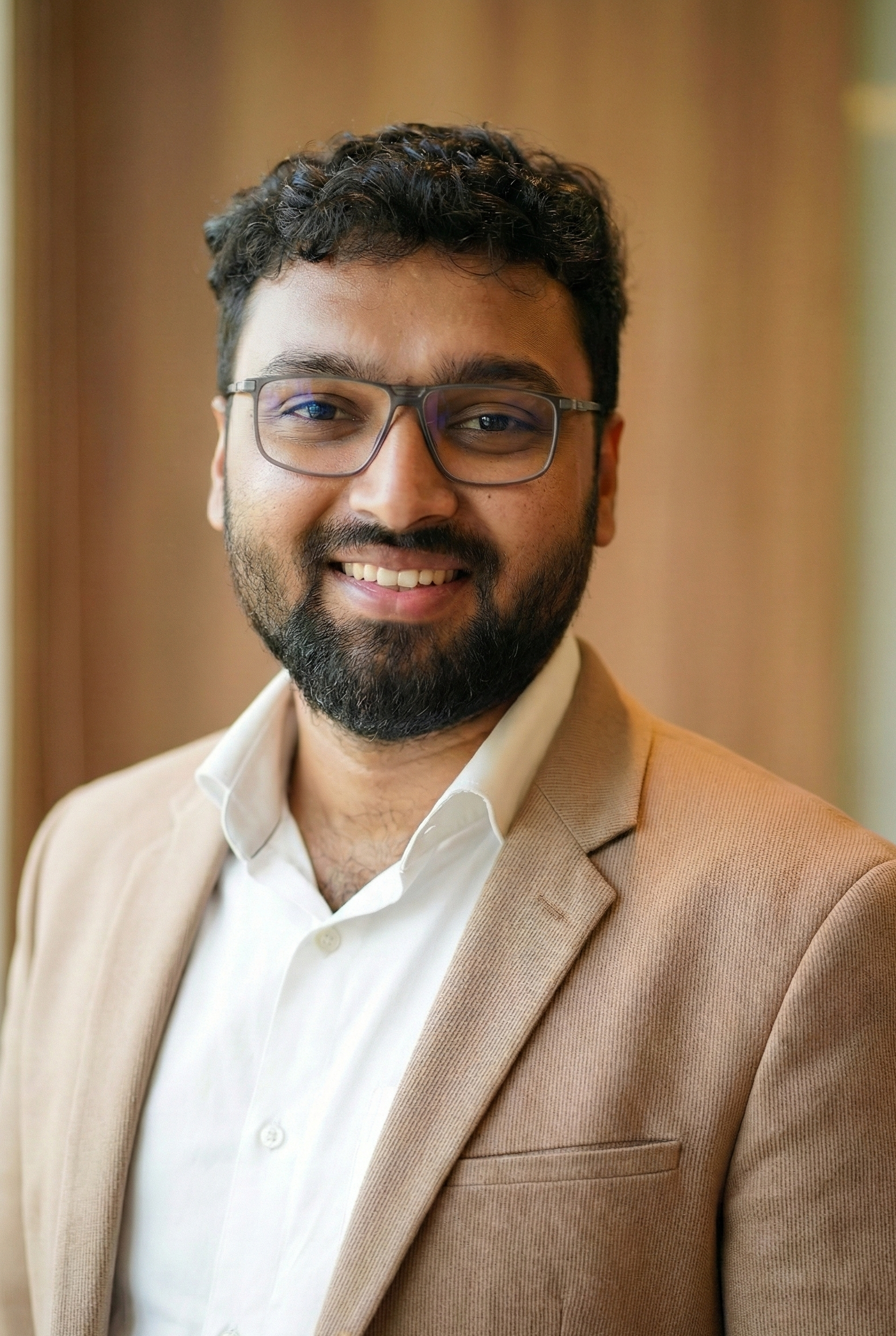 A smiling man with glasses, beard, and mustache, wearing a beige suit jacket and white shirt, standing indoors near a window.