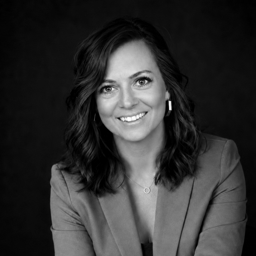 Black and white portrait of a woman with wavy hair, wearing earrings, a necklace, and a blazer, smiling at the camera.