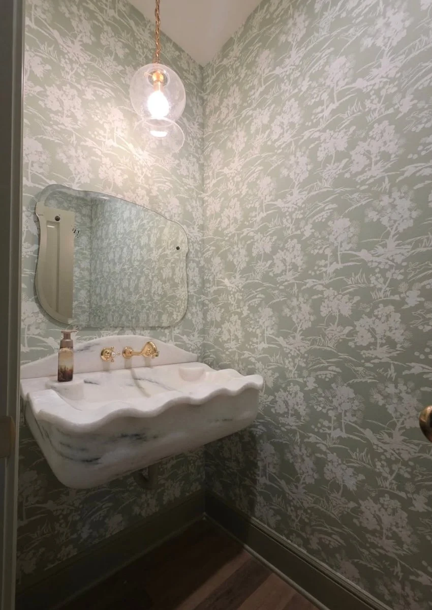 Small vintage bathroom with a marble sink and an ornate mirror above it, floral wallpaper, and a hanging light fixture.