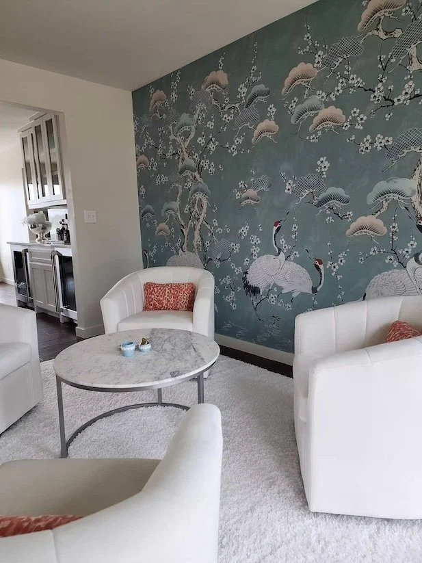 Living room with white armchairs, a marble-top coffee table with small bowls, and a large decorative wallpaper mural featuring cranes, cherry blossoms, and flowing water in shades of blue, gray, and white.
