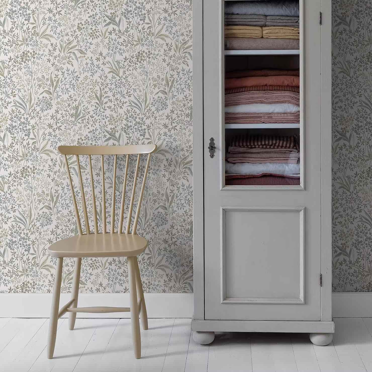 Light wooden chair next to a white cabinet with folded towels, set against a floral-patterned wallpaper background.