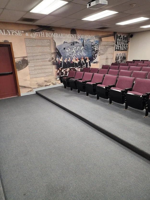 Empty auditorium with maroon and black seats, a wall display about the 436th Bombardment Squadron, and a projector on the ceiling.