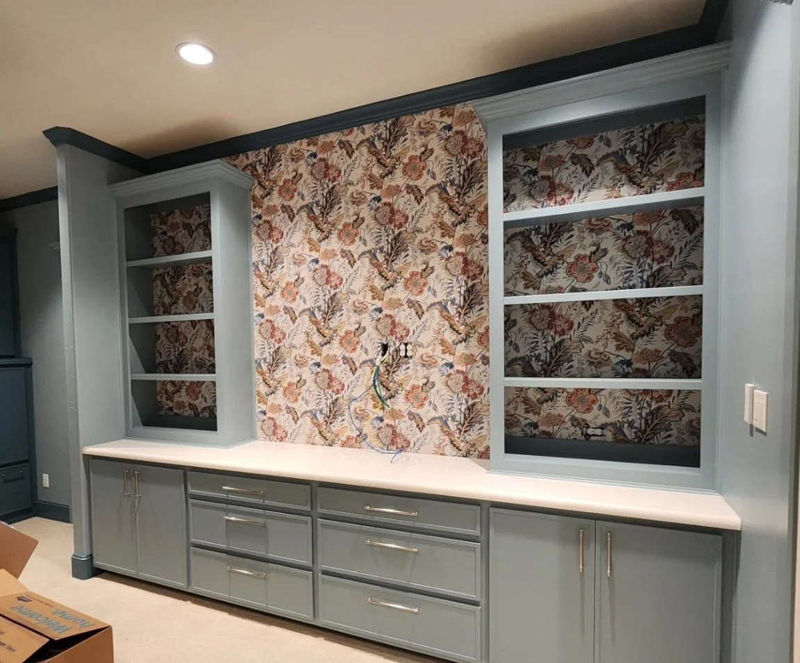 Custom built light blue cabinet with open shelves on either side, set against floral wallpapered wall, with electrical wiring visible in the center, in a room under renovation.
