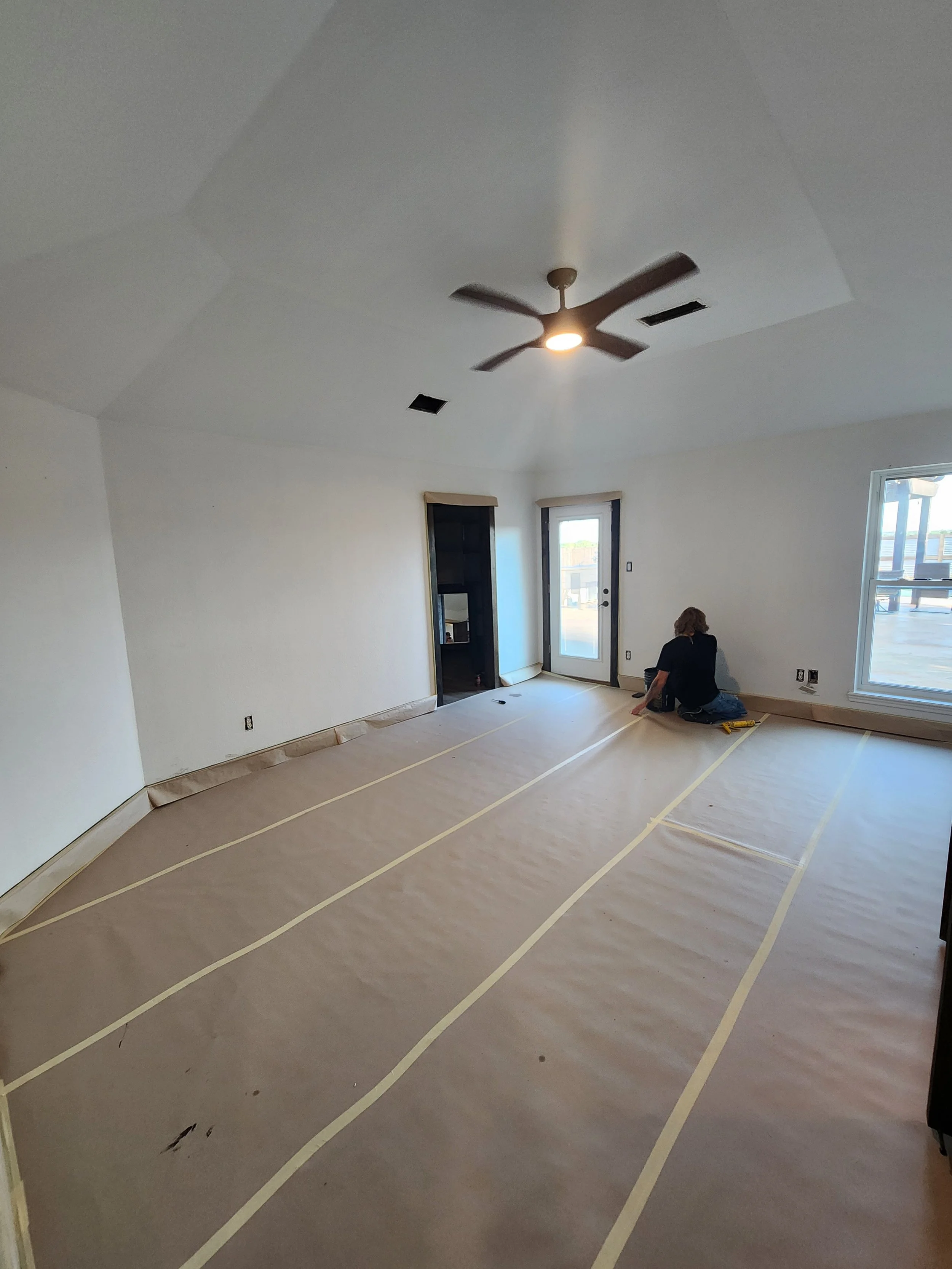 A room under renovation with a person kneeling on the floor, which is covered with paper and masking tape, near a door and a window, with a ceiling fan and an overhead light.