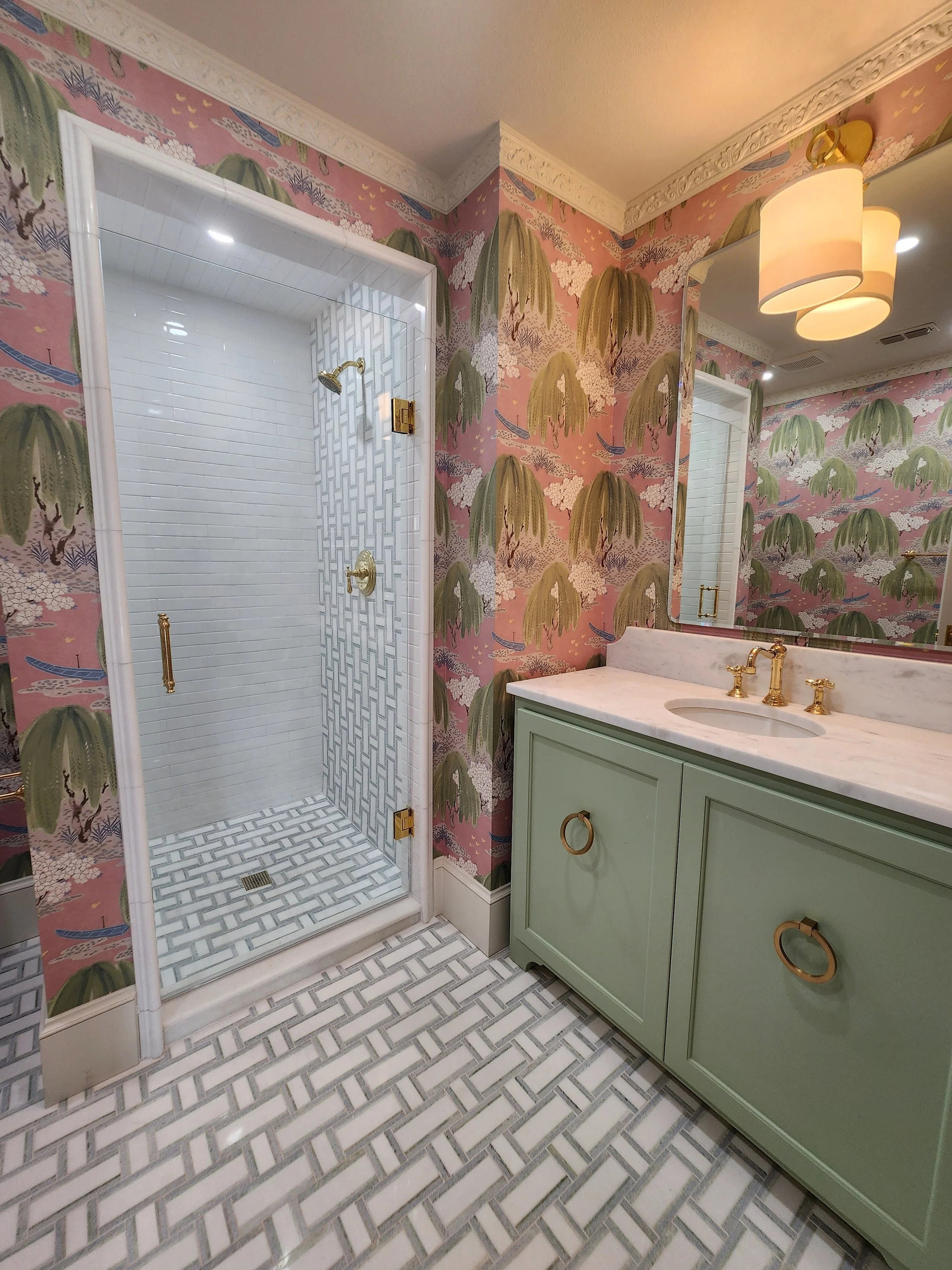 Bathroom with pink floral wallpaper, green vanity with gold hardware, large mirror, and a glass shower with white subway tiles and geometric patterned floor and shower walls.