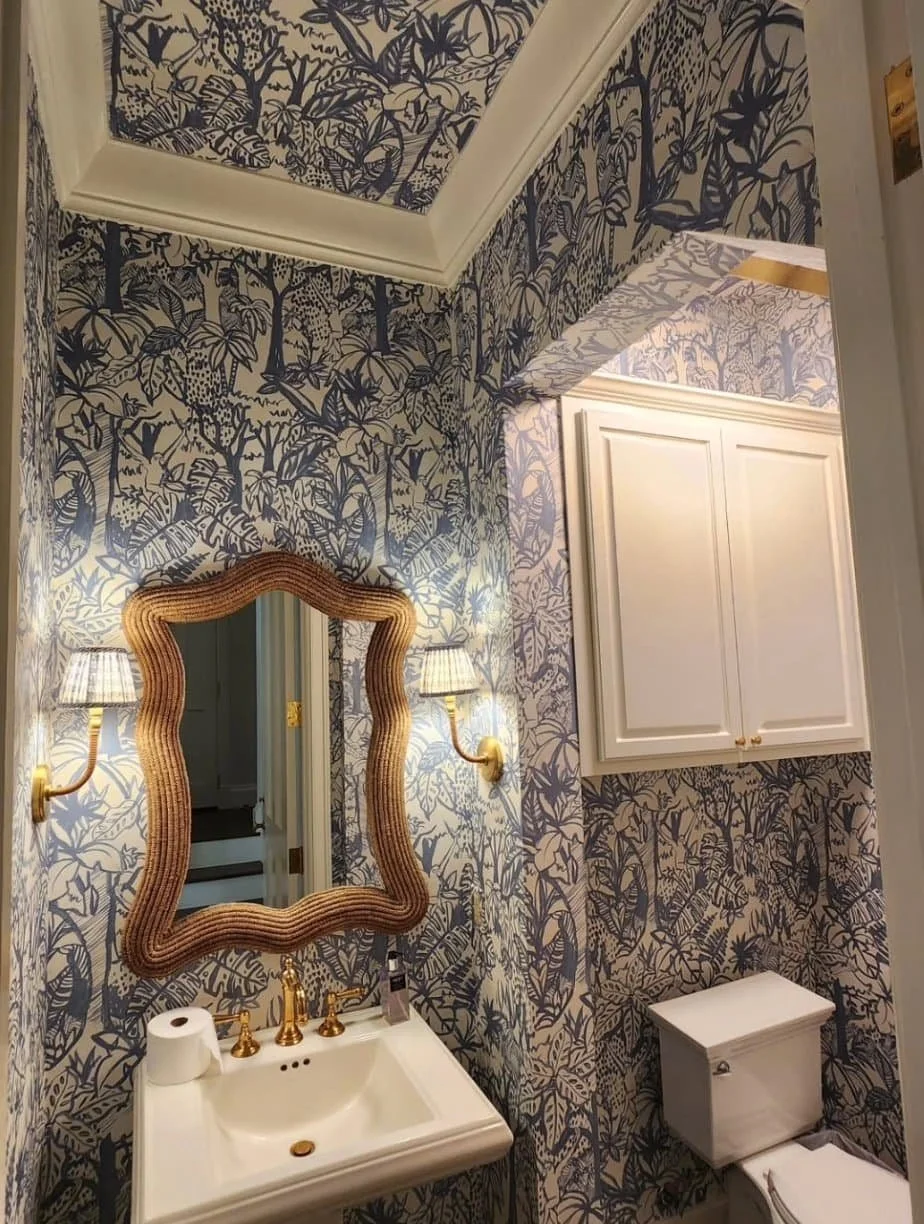 A small bathroom with blue and white botanical wallpaper, a wavy framed mirror above a white sink with gold fixtures, and wall-mounted lamps on either side of the mirror. There is a toilet below a white cabinet on the right side.