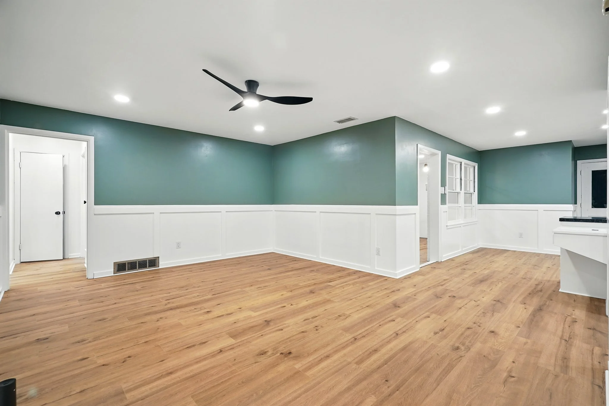 Empty living room with wooden floors, green walls with white wainscoting, ceiling fan, recessed lighting, and a window.