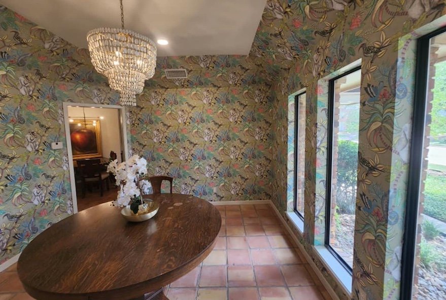 A dining room with floral wallpaper, a wooden oval table with a white flower arrangement, a chandelier, and three large windows showing greenery outside.