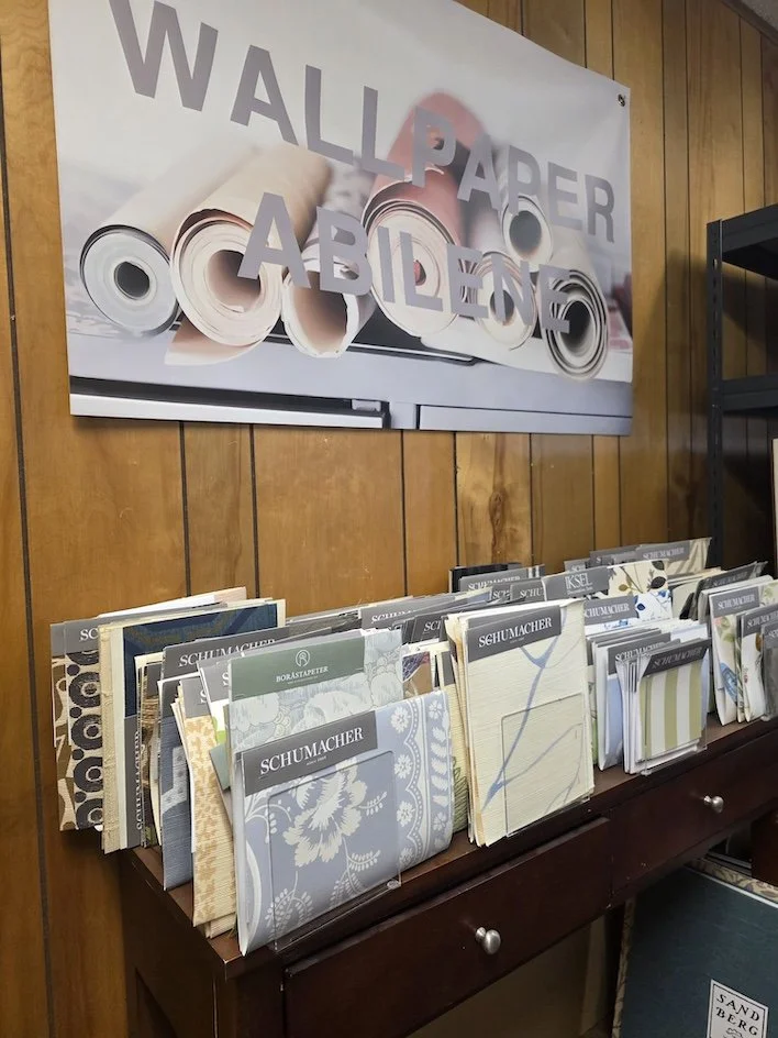Display of wallpaper samples in a store with a sign that reads "WALLPAPER AVAILABLE" in the background.