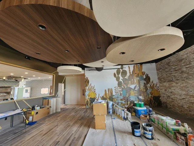 Restaurant under construction with wood flooring, large ceiling panels, mural wall, supplies, and commercial wallpaper installation in progress.
