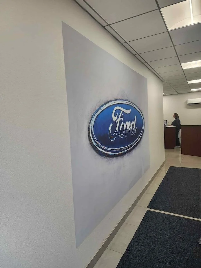 Interior of a Ford dealership with a large Ford logo on the wall and a receptionist desk in the background.