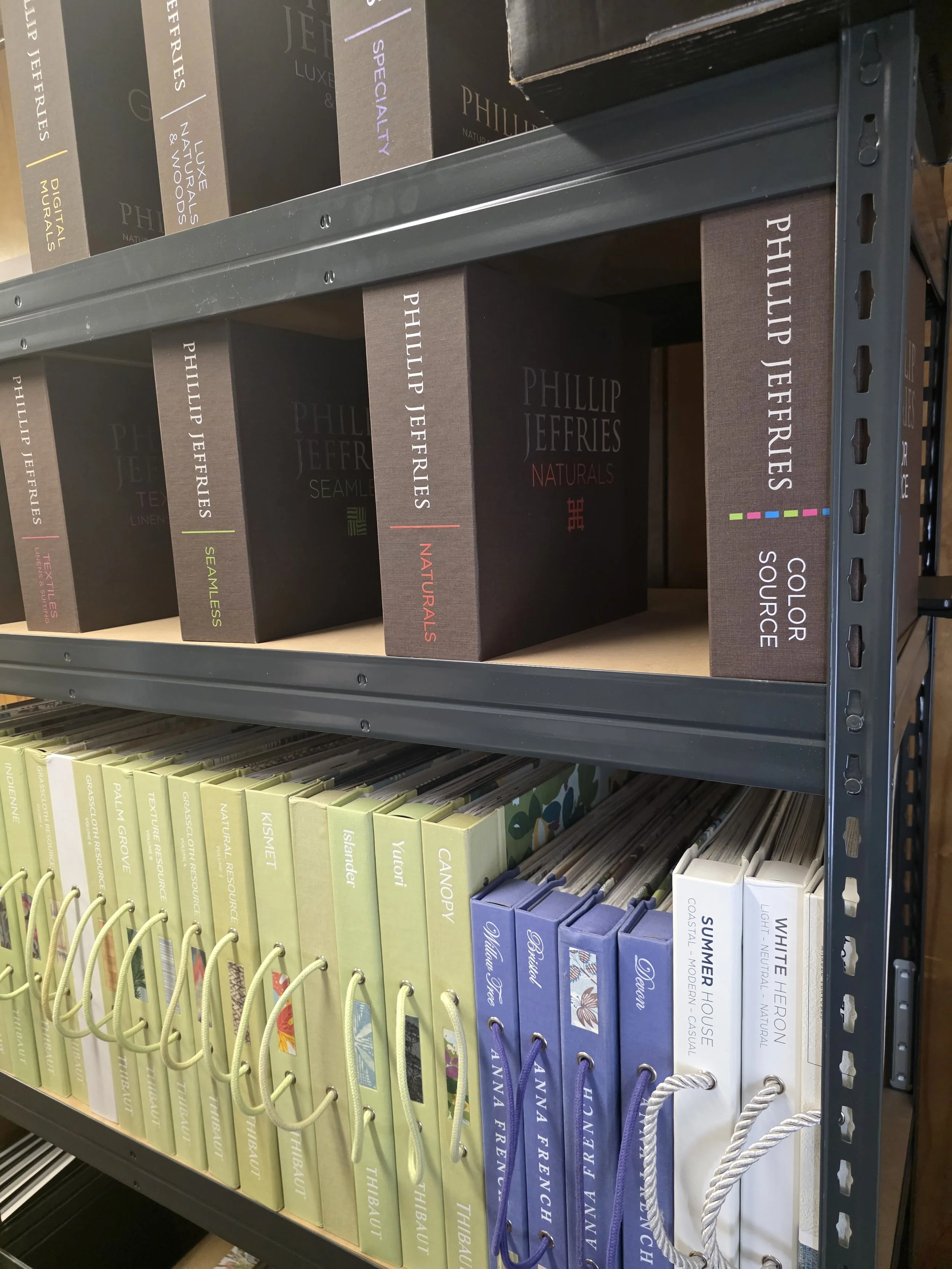 Shelf with books and boxes of Philip Jeffries products, including 'Naturals,' 'Seamless,' and 'Textiles,' with a lower row of colorful fabric sample folders.