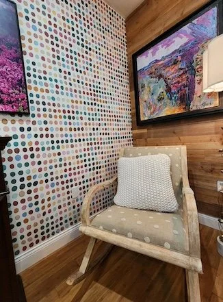 A cozy corner with a patterned polka dot wall, wooden wall paneling, and two colorful abstract paintings, featuring a white armchair with a cushion.