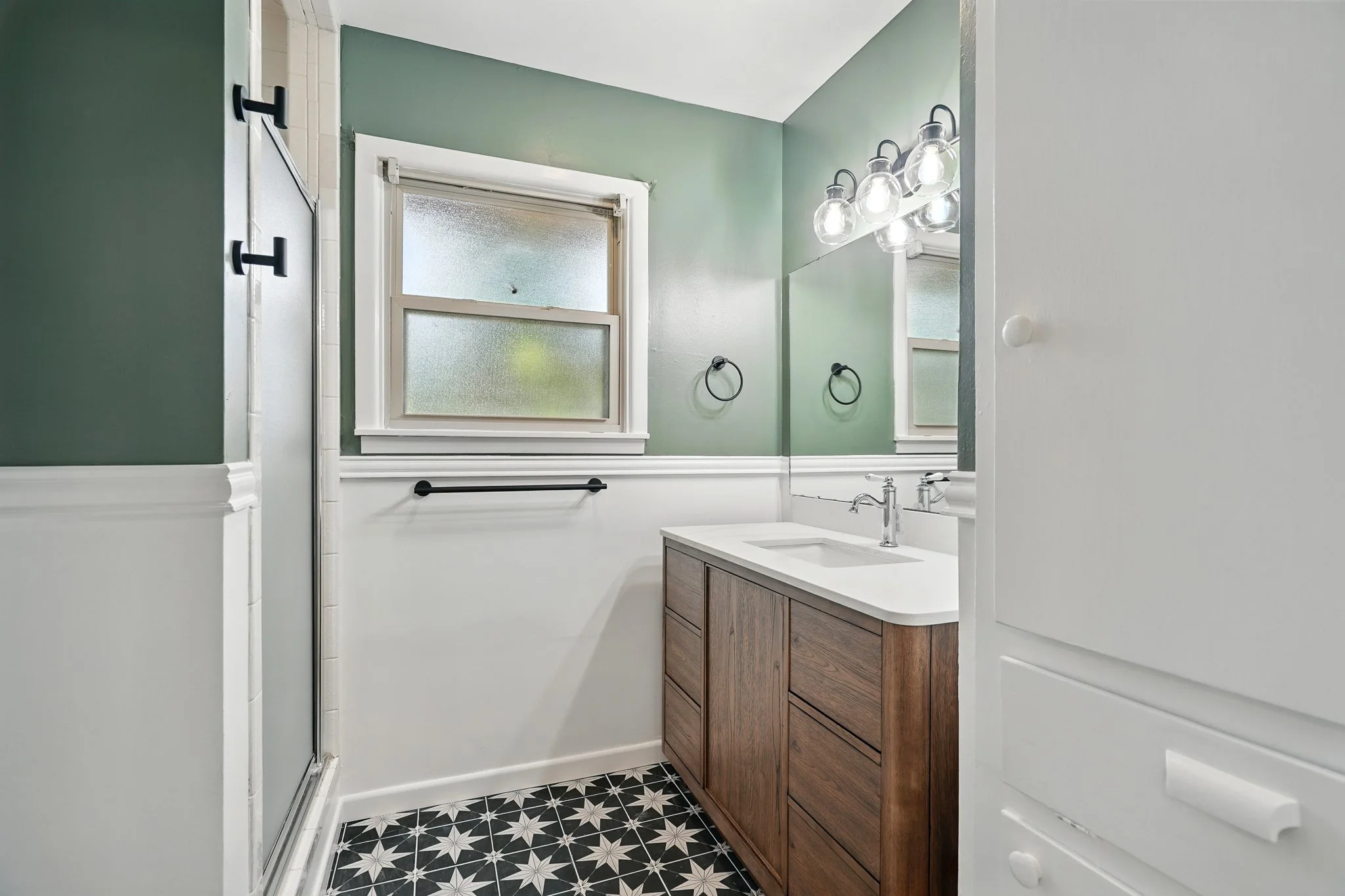 Small bathroom with a green and white color scheme, a frosted window, a wooden vanity with a white sink, and decorative black and white floor tiles.