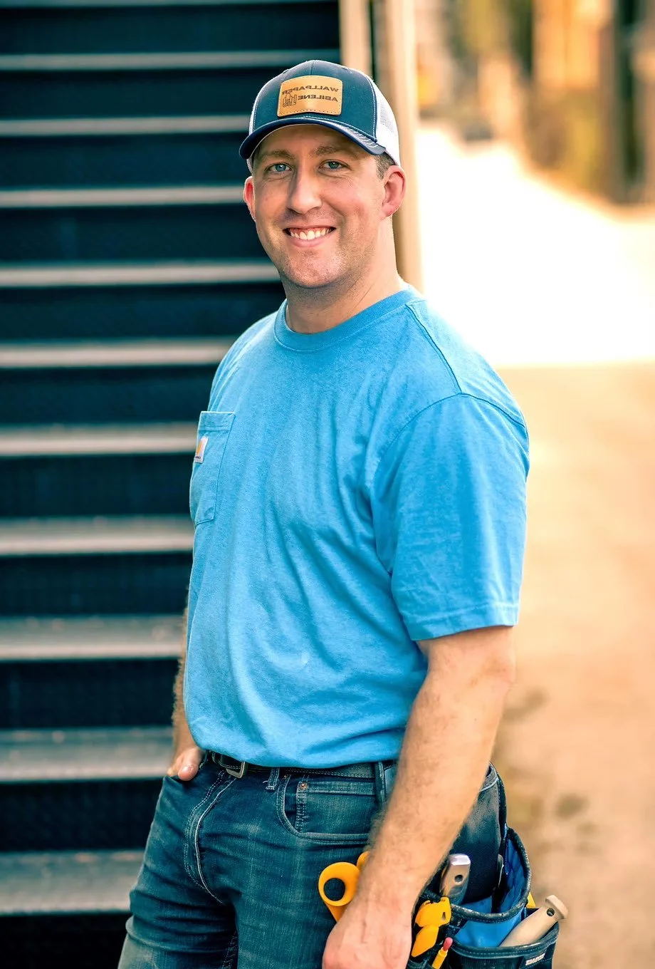 A smiling man in a blue t-shirt and cap with a tool belt standing outdoors near a staircase with sunlight in the background.