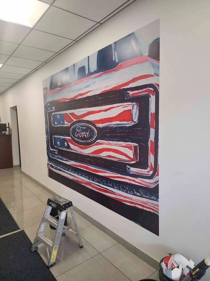 A wall mural of a Ford vehicle grille with an American flag pattern background in a commercial space. A small stepladder and some tools are in front of the wall.